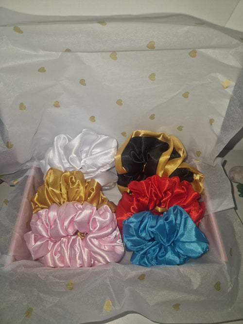 Six satin hair scrunchies in black, gold, red, blue, white, and pink, arranged inside a pink gift box lined with white tissue paper decorated with small gold hearts.