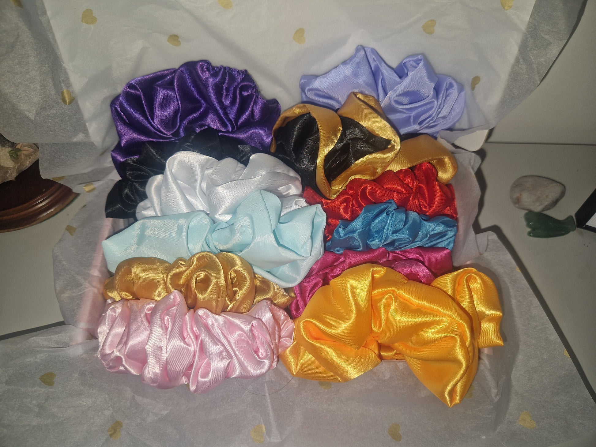 A collection of satin hair scrunchies in multiple colors, including purple, black, white, light blue, red, royal blue, pink, and metallic gold, arranged on white tissue paper with gold hearts.