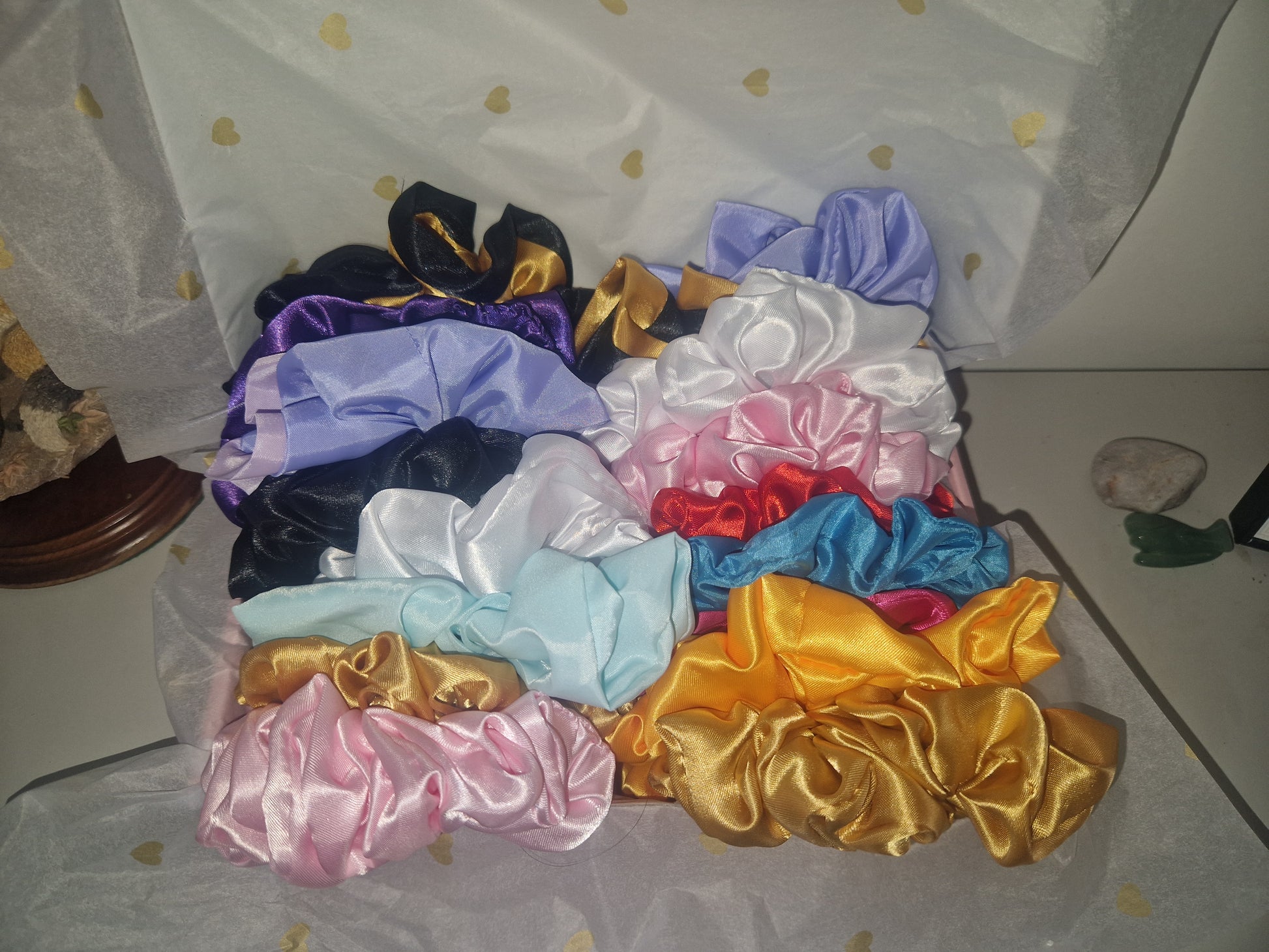 A collection of satin  hair scrunchies in multiple colors, including purple, black, white, light blue, red, royal blue, pink, and metallic gold, arranged on white tissue paper with gold dots.