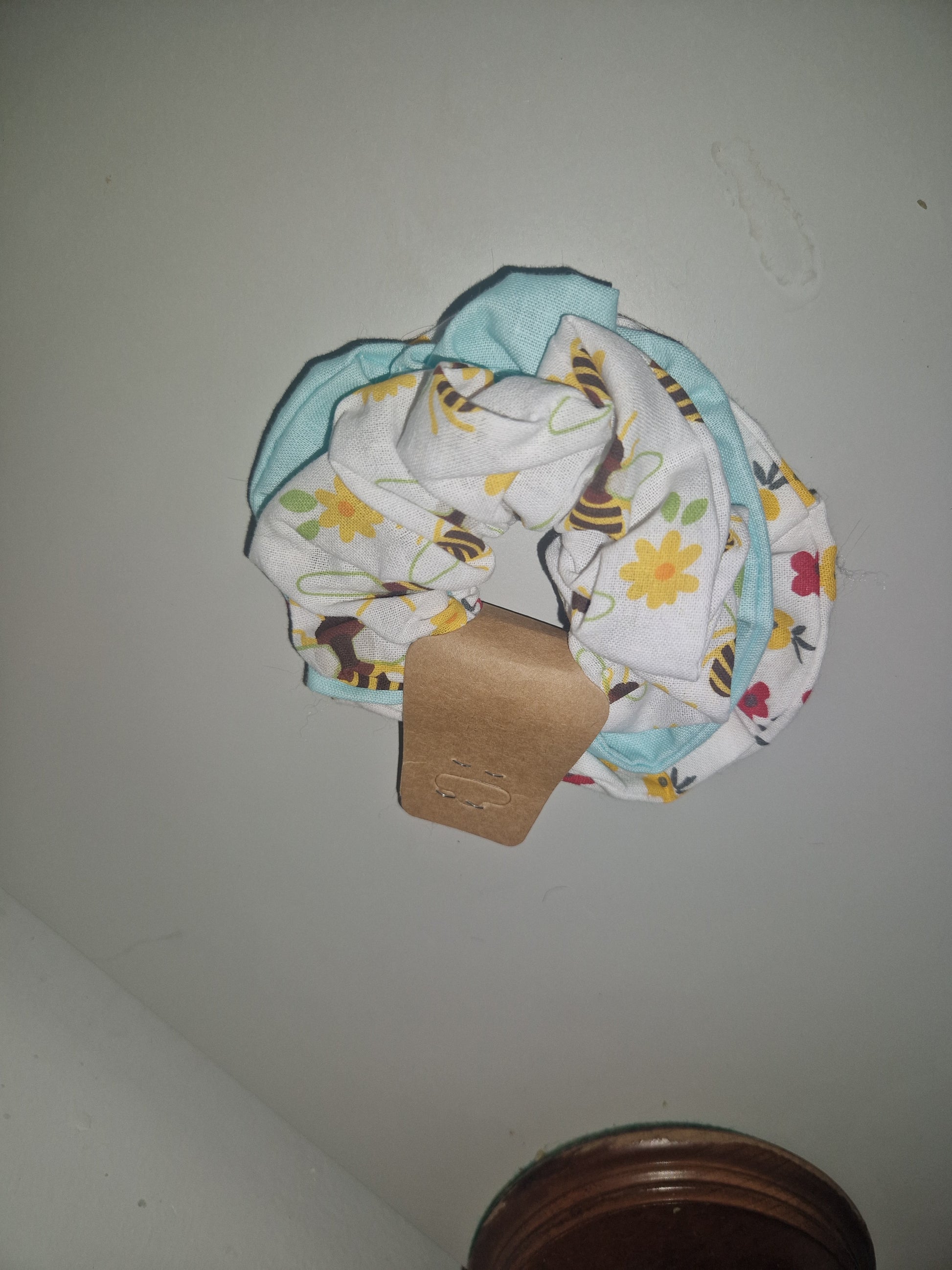 A folded baby fabric accessory, likely a burp cloth or muslin, featuring a white background with a yellow flower and bee print, layered with a light blue fabric and a brown paper tag, set against a plain gray wall.