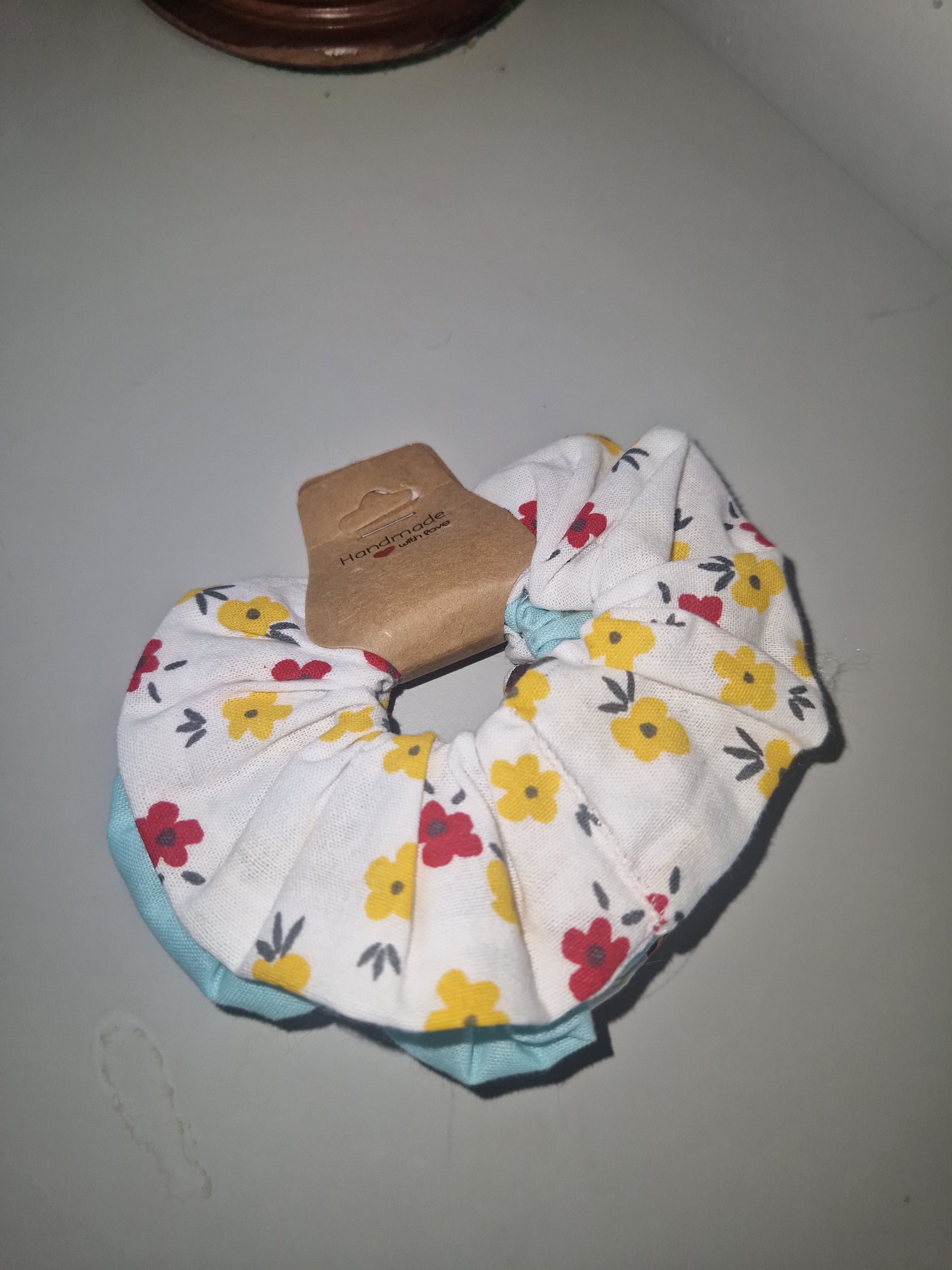 A fabric hair scrunchie with a white background and a colorful floral print featuring yellow, red, and dark green flowers, finished with a light blue edge trim and a brown cardboard hanger.