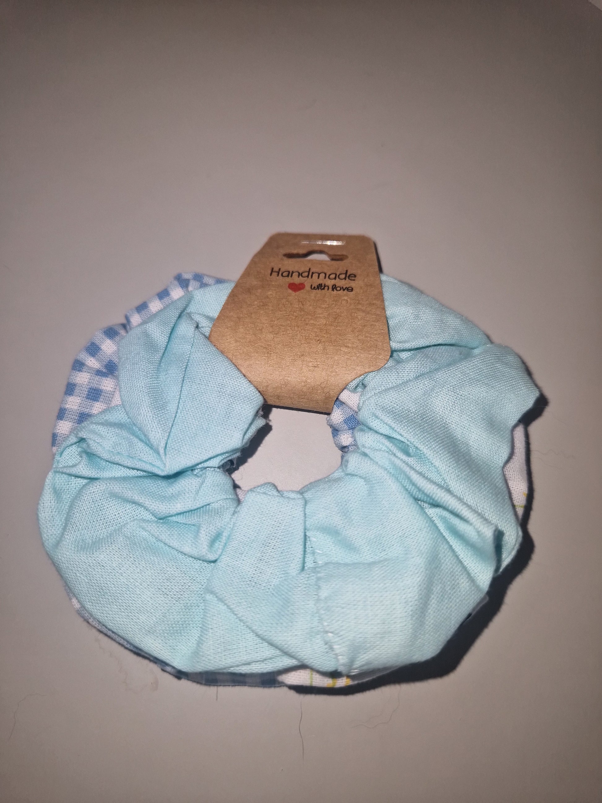A light blue fabric hair scrunchie with a brown cardboard tag attached, photographed on a plain beige background.