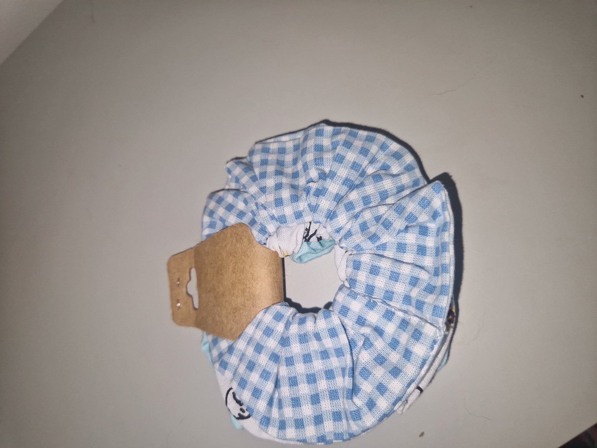 A fabric hair scrunchie with a blue and white gingham check pattern, shown with its brown cardboard hanger tag attached, set against a plain light gray background.