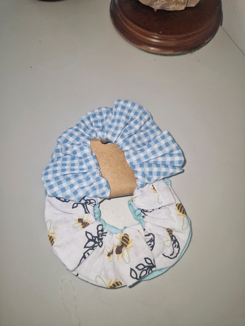 A handmade cotton hair scrunchie featuring a blue and white gingham check pattern on one side and a white fabric with a bee print on the other, displayed on a neutral surface.