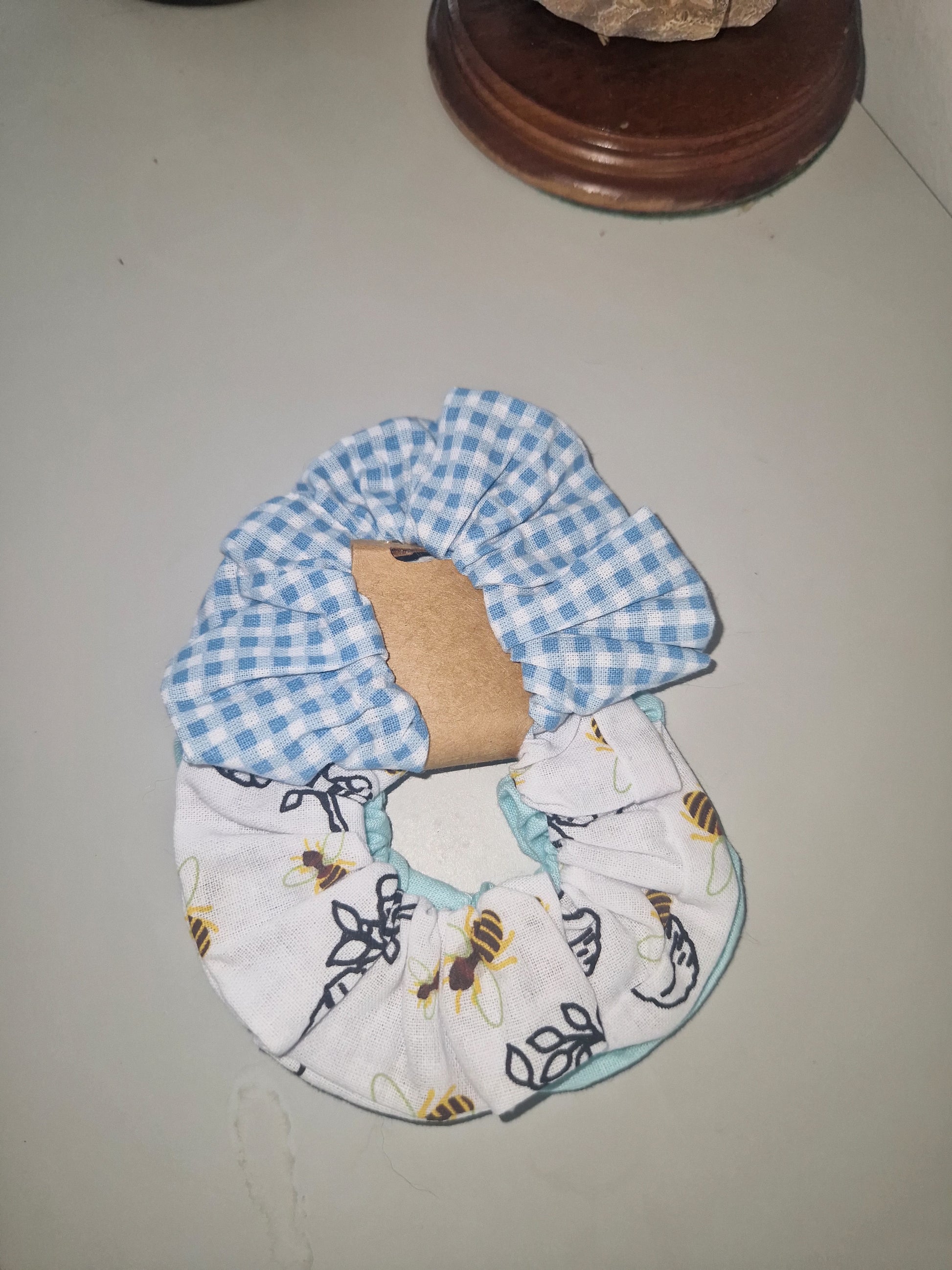 A handmade cotton hair scrunchie featuring a blue and white gingham check pattern on one side and a white fabric with a bee print on the other, displayed on a neutral surface.