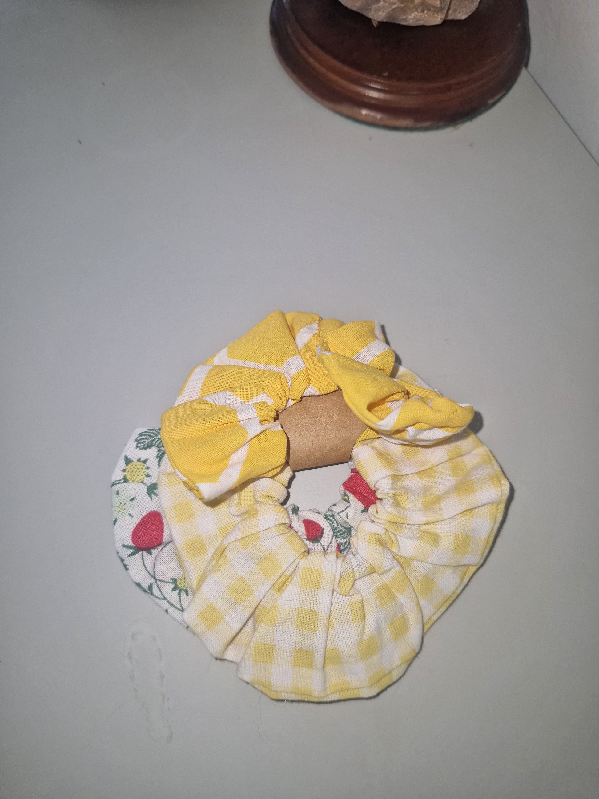 A fabric hair scrunchie with a layered design, featuring a yellow and white gingham check pattern on the top half and a white bottom half with a strawberry and green leaf floral print. A brown cardboard tube is tucked inside the center of the scrunchie, which is photographed on a plain light gray surface.
