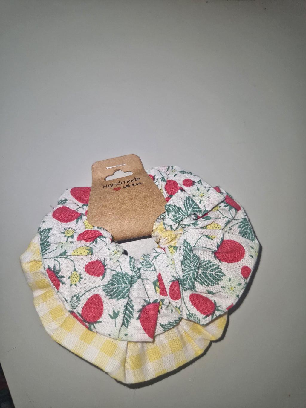 A fabric hair scrunchie with a strawberry and leaf print on a white background, layered with a yellow and white gingham check pattern, attached to a brown cardboard hanger.