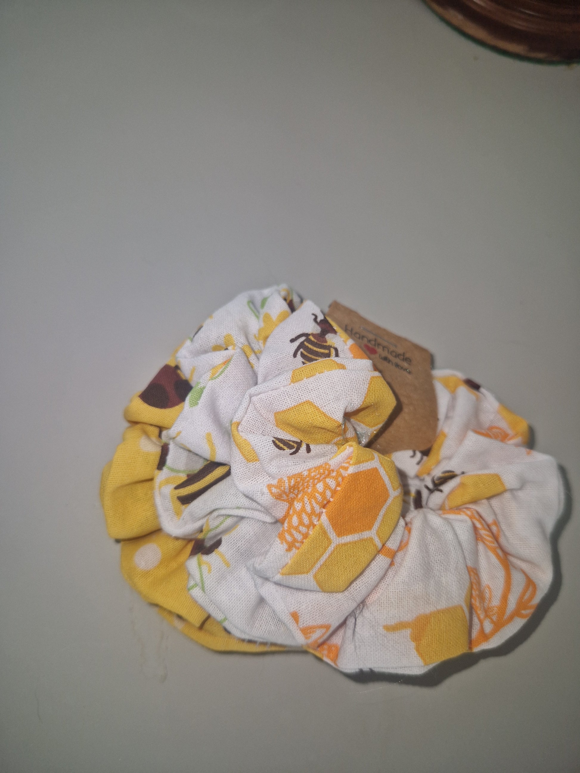 A fabric hair scrunchie with a honeycomb and bee pattern in yellow, orange, and white, featuring a brown paper tag attached, set against a plain light gray background.