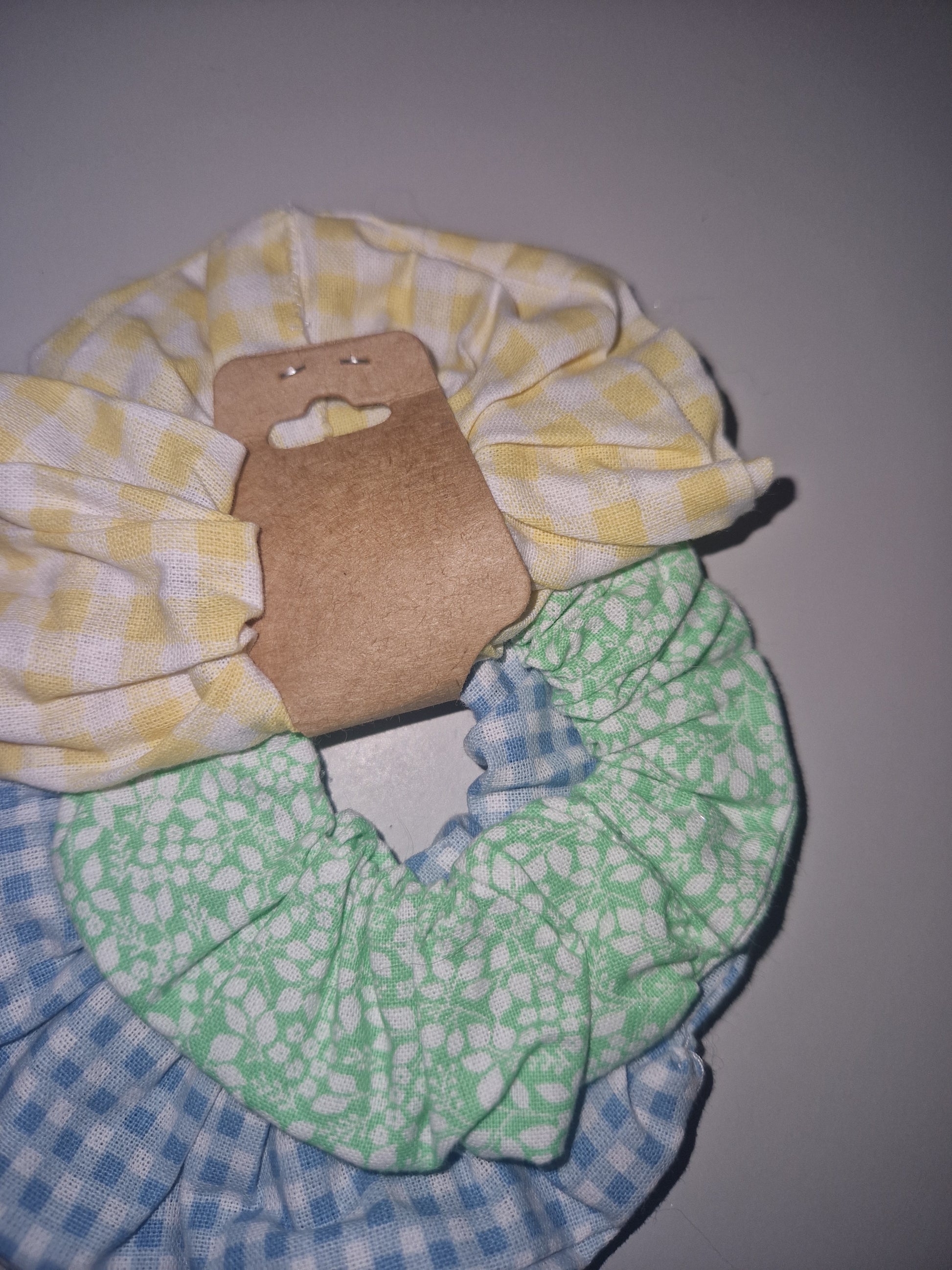 Three fabric hair scrunchies with different patterns (yellow and white gingham, mint green and white floral, and blue and white gingham) arranged together on a neutral gray background, with a cardboard hanger tag attached.