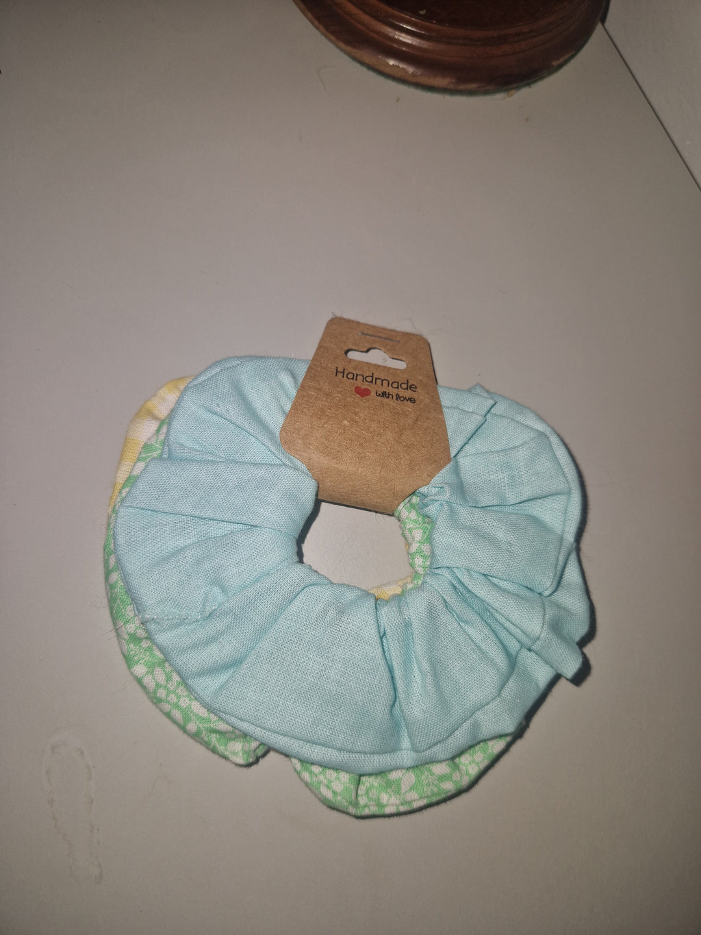 A fabric hair scrunchie in light blue with a green and white patterned lining, attached to a brown cardboard hang tag, shown on a plain light gray surface.