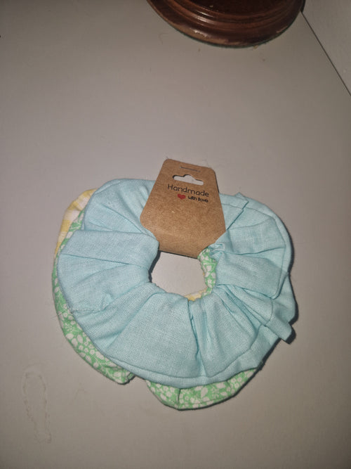A fabric hair scrunchie in light blue with a green and white patterned lining, attached to a brown cardboard hang tag, shown on a plain light gray surface.