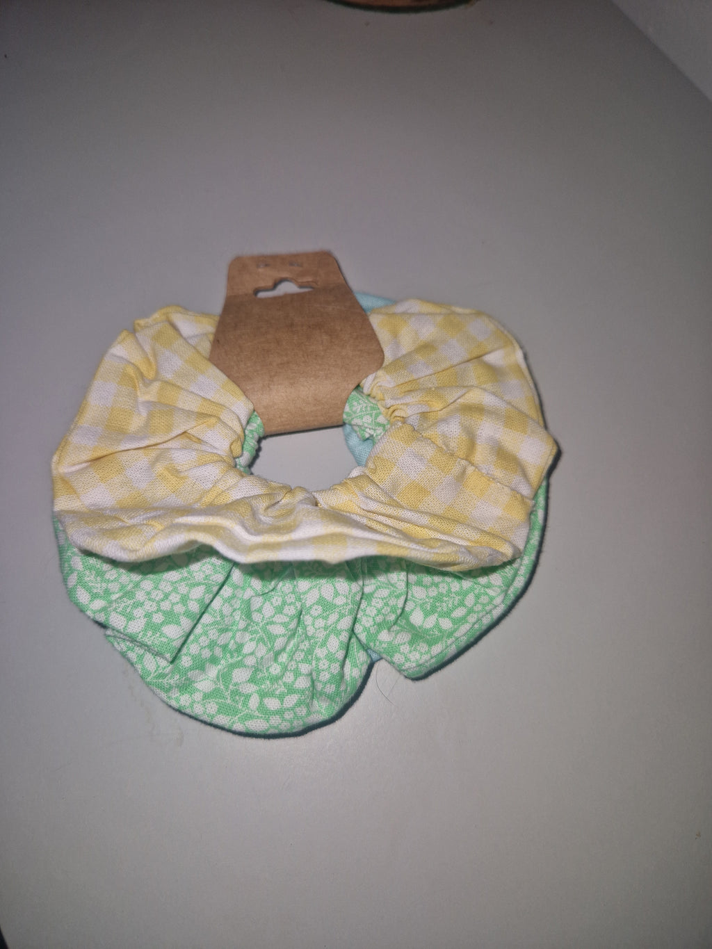 Two fabric hair scrunchies displayed on a cardboard hanger against a plain light gray wall. One scrunchie is yellow and white gingham, and the other is mint green and white floral.