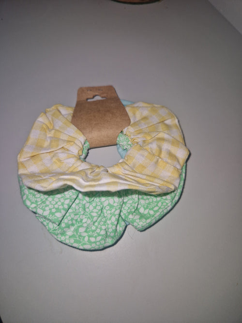 Two fabric hair scrunchies displayed on a cardboard hanger against a plain light gray wall. One scrunchie is yellow and white gingham, and the other is mint green and white floral.