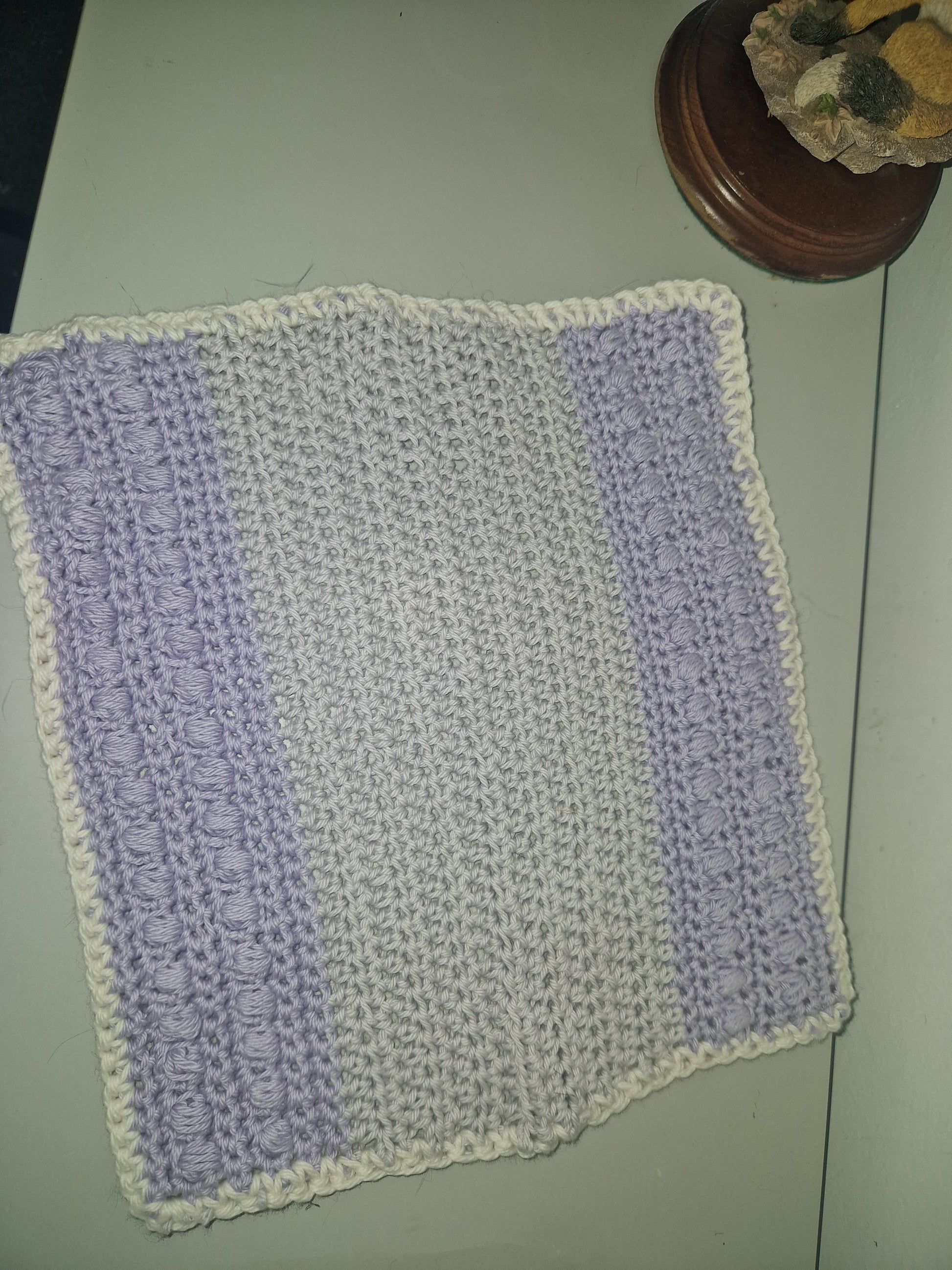 A handmade crocheted flannel with horizontal stripes of lavender purple and off-white, finished with a matching off-white border, laid flat on a light-colored surface.