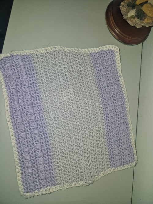A handmade crocheted flannel with horizontal stripes of lavender purple and off-white, finished with a matching off-white border, laid flat on a light-colored surface.