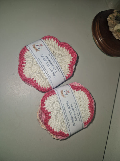 Two handmade crocheted face scrubbers with pink and white yarn, wrapped in white paper bands labeled 'HANDMADE FACE SCRUBBIES', placed on a light gray surface.