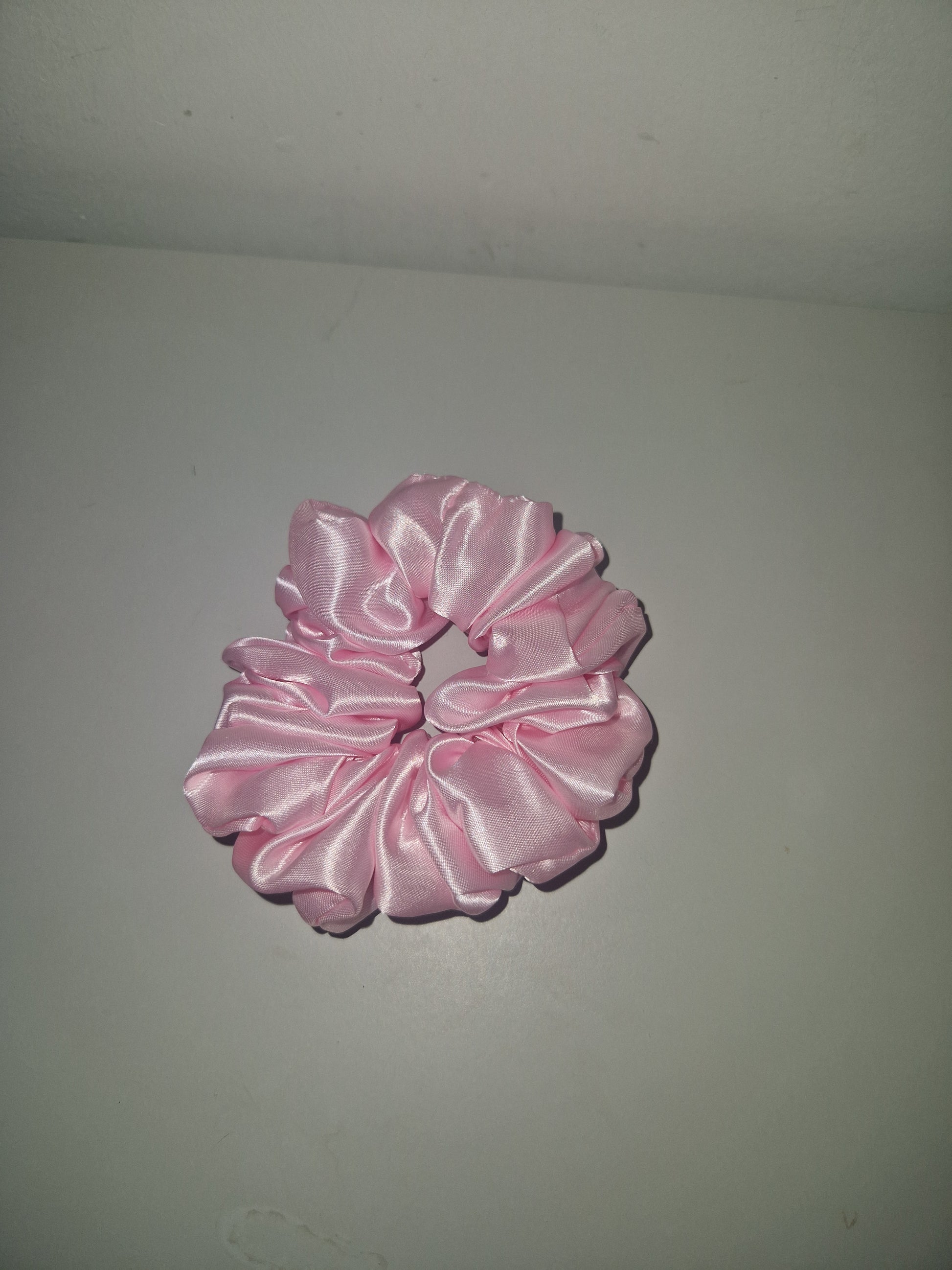 A light pink satin hair scrunchie displayed against a neutral gray background.