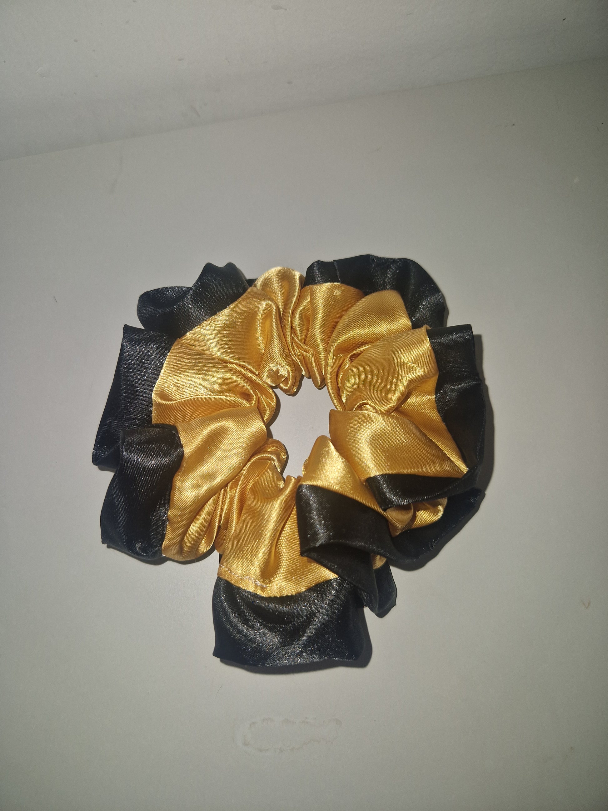 A satin hair scrunchie with alternating ruched panels of black and metallic gold, shown against a plain off-white background.