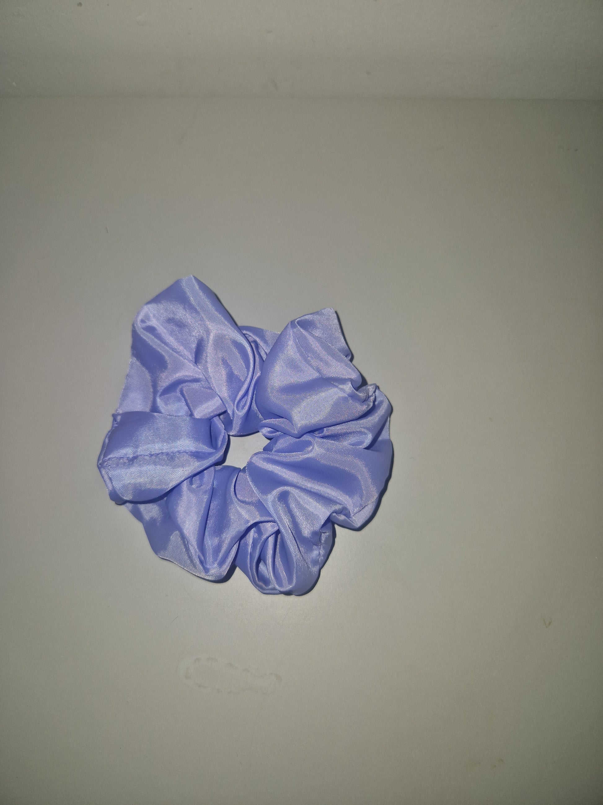 A light purple satin hair scrunchie displayed against a plain, light gray background.