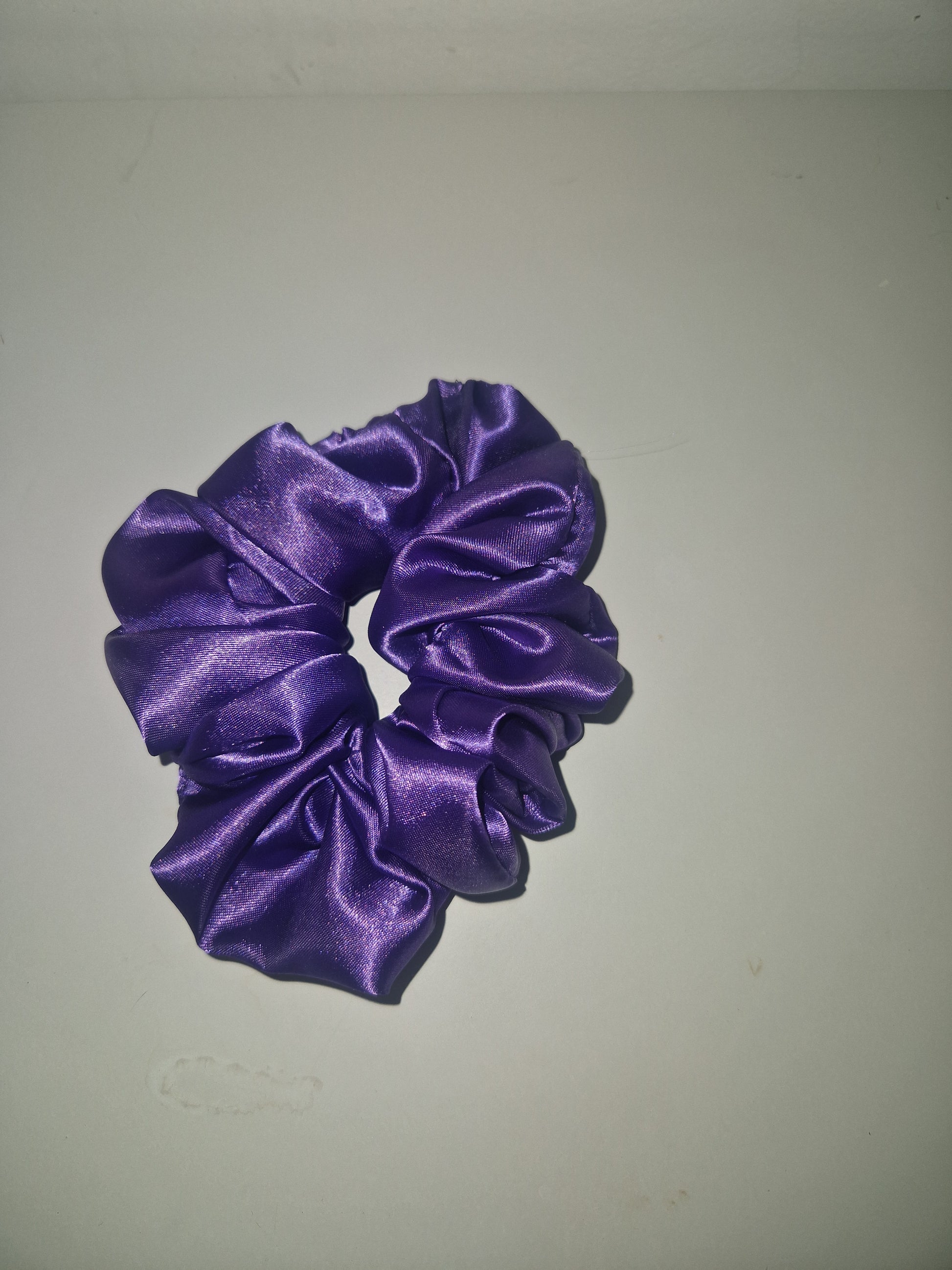 A purple satin scrunchie with a shiny, lustrous finish, photographed against a plain off-white background.