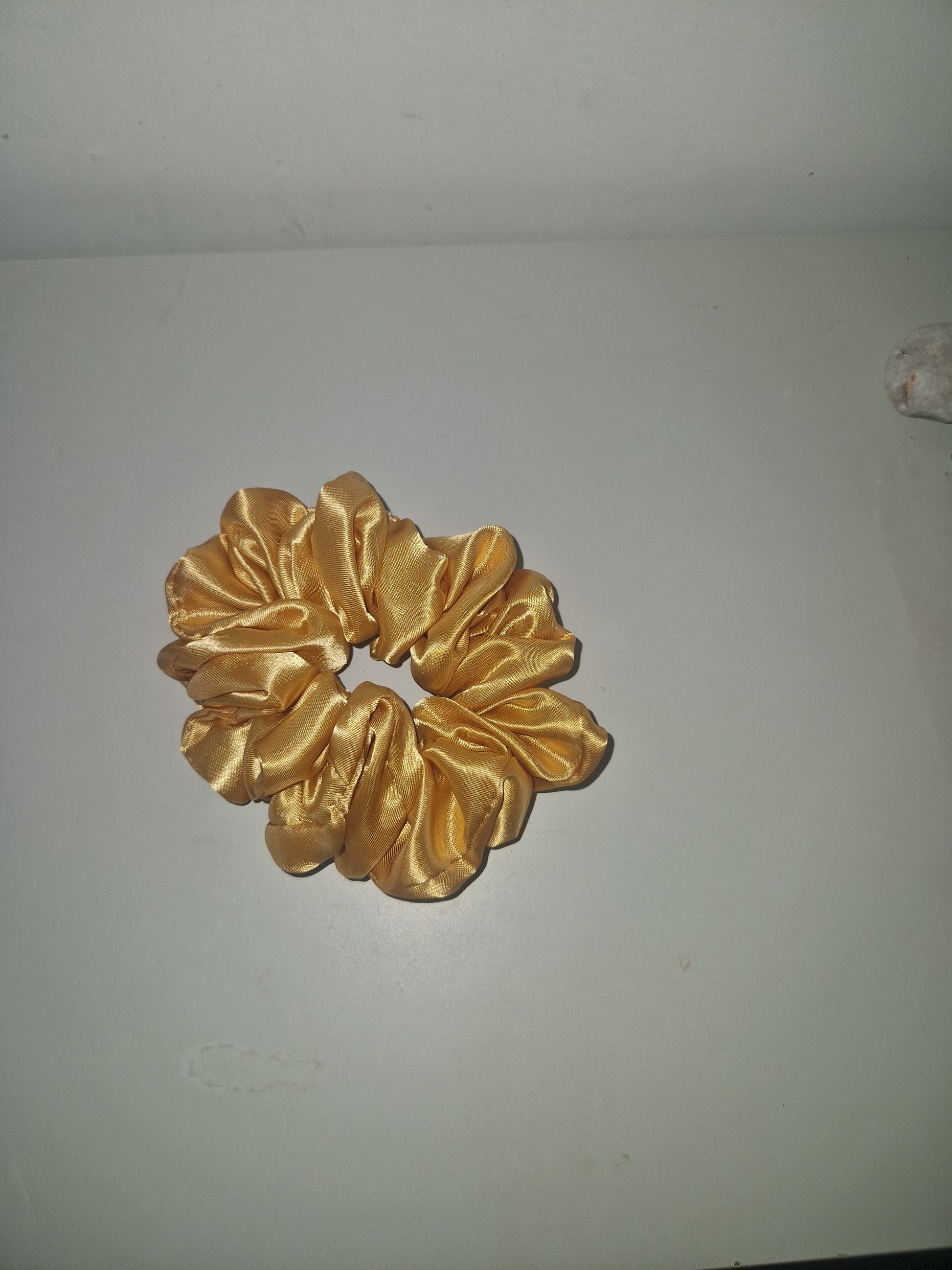 A shiny gold satin scrunchie hair tie displayed against a plain off-white wall.