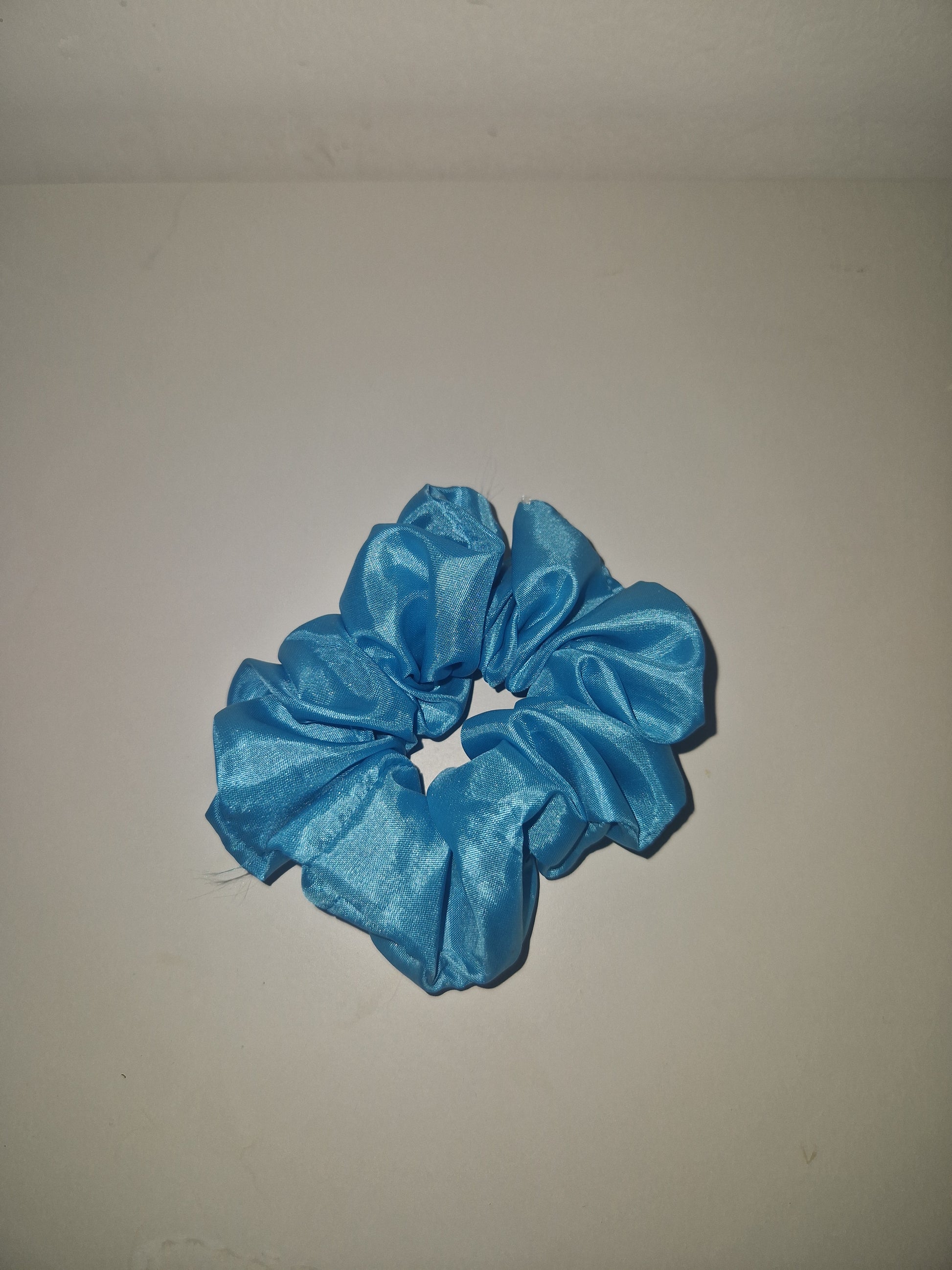 A single bright blue satin hair scrunchie displayed against a plain, off-white background.