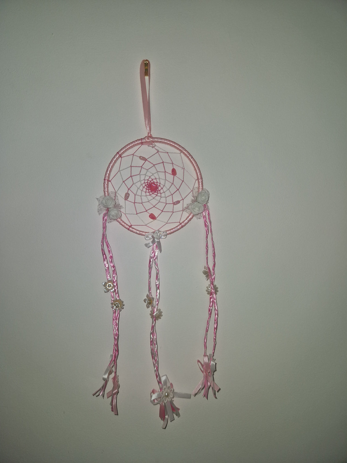 A pink dreamcatcher hair clip with a circular web design, adorned with small white flowers and dangling pink ribbon tassels, shown on a plain light gray background.