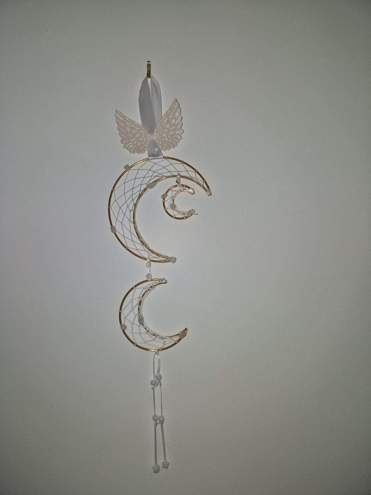 A decorative hair accessory featuring a white feather, a white lace wing, and three gold crescent moon dreamcatcher-style loops with white webbing, shown against a plain light gray background.