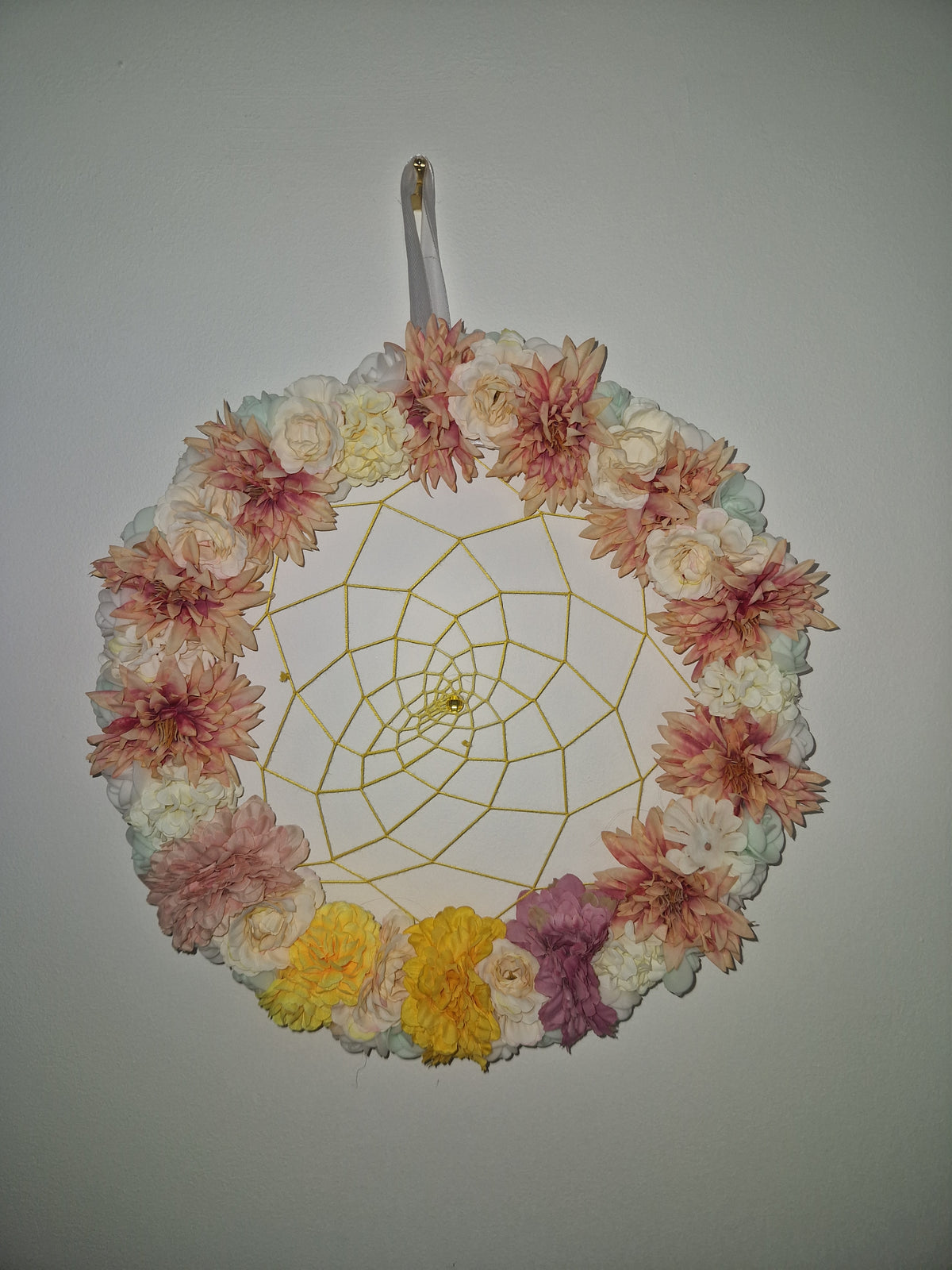 A circular decorative fan with a white handle, decorated with a ring of dried flowers in pink, peach, yellow, and white, and featuring a gold-colored web design in the center, shown against a plain light gray background.
