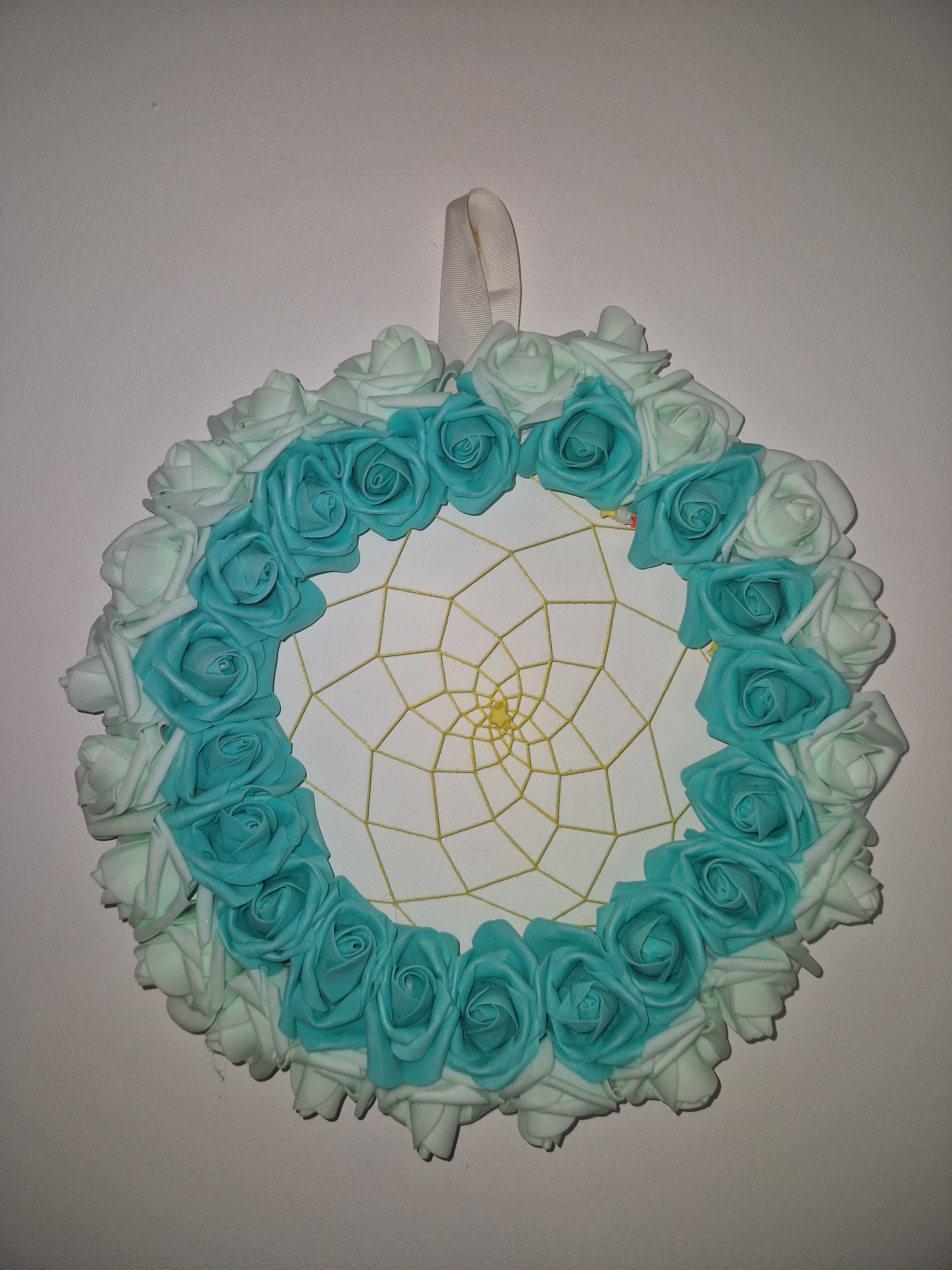 A circular decorative floral wreath made of layered fabric roses in aqua blue and mint green, with a white center and a gold spiderweb design, shown against a plain light background.