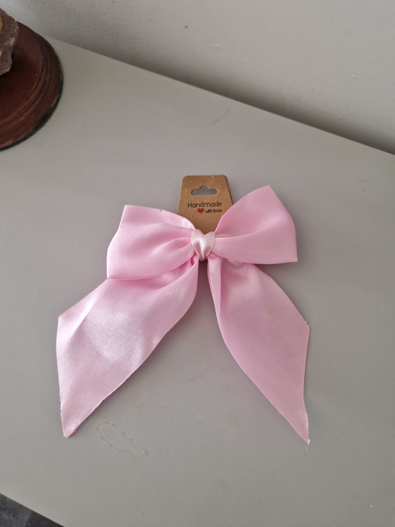 A light pink satin bow with a small cardboard tag attached, resting on a neutral gray surface.