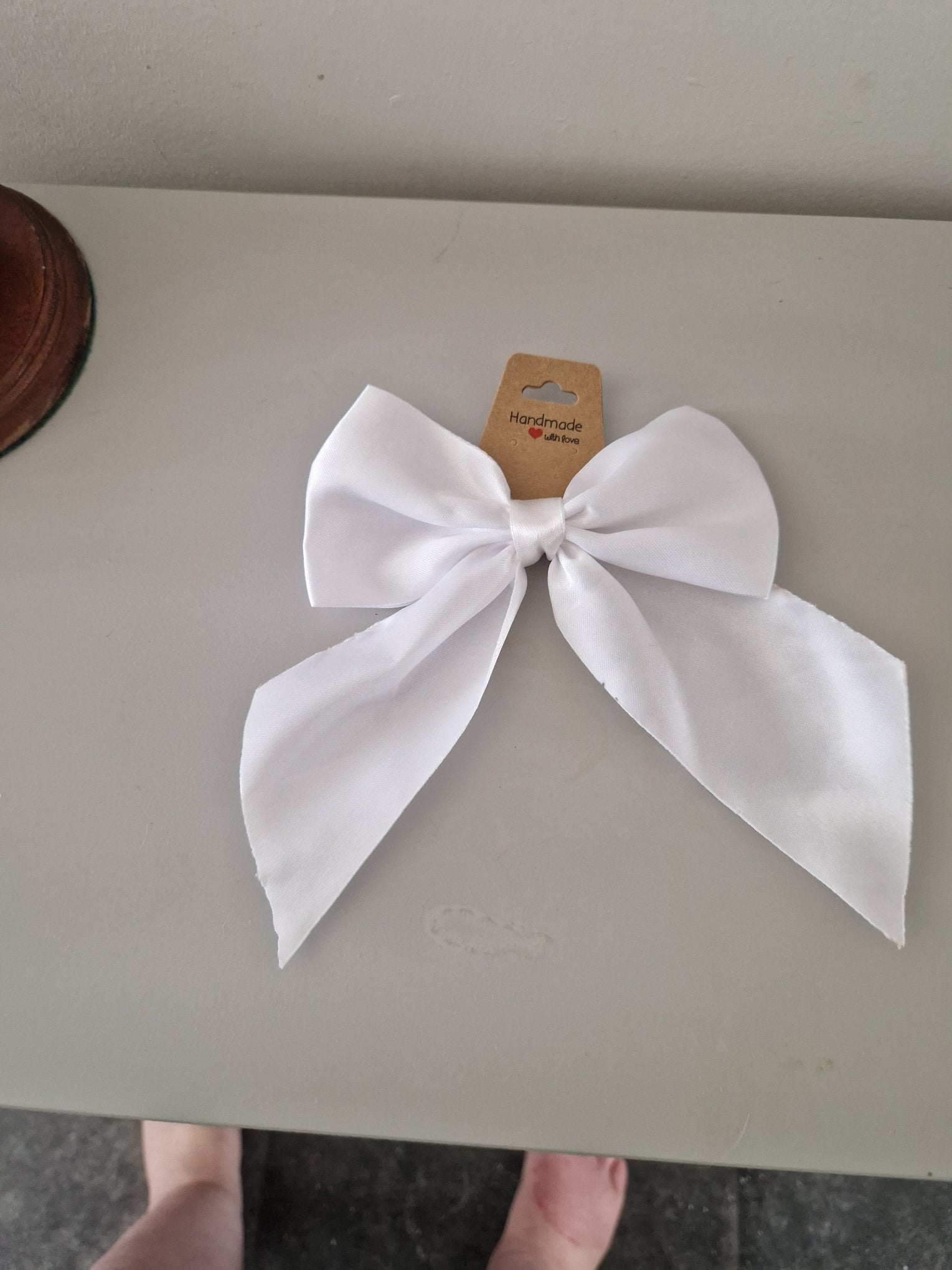 A white satin bow with a small tan paper tag, placed on a light gray surface.