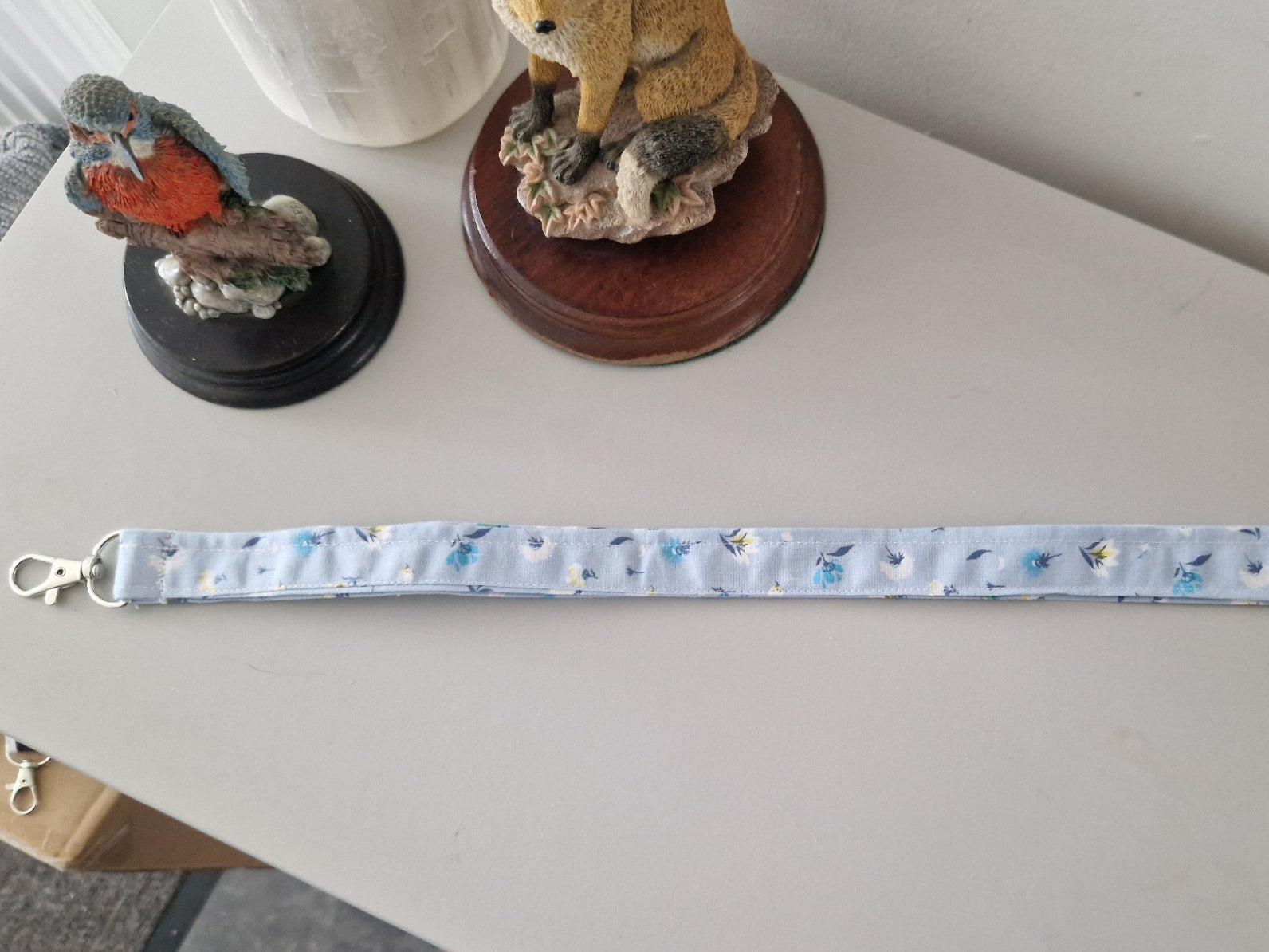 A light blue fabric strap with a small blue and white pattern, featuring silver metal lobster clasps on both ends, laid out on a light gray table. In the background, there are decorative figurines on a shelf, and a foot is visible at the bottom edge.
