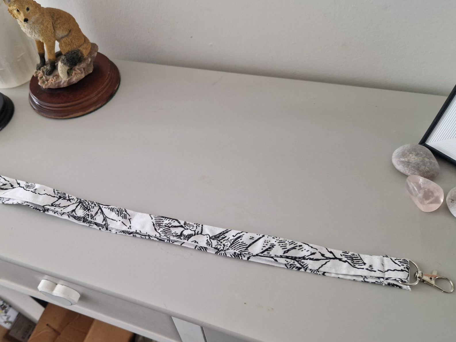 A long fabric strap or lanyard with a black and white botanical leaf pattern, laid out on a light gray dresser. The strap has metal lobster clasps at both ends and is photographed in a home setting with decorative figurines and crystals on the dresser.