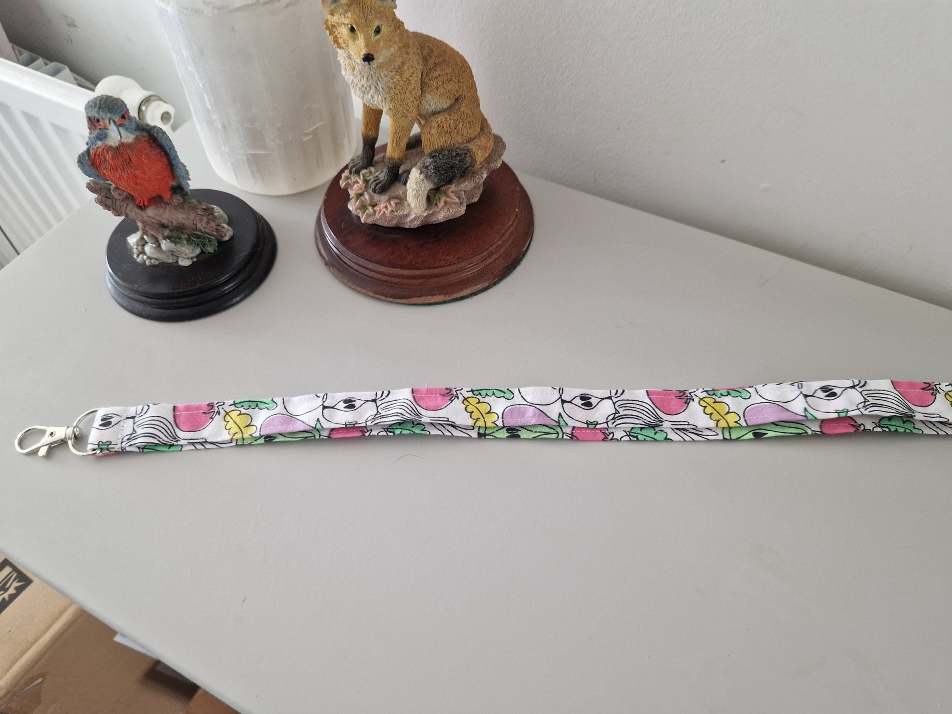 A fabric lanyard strap with a colorful cartoon fruit pattern and a silver metal lobster clasp, laid out on a light gray tabletop with decorative figurines in the background.