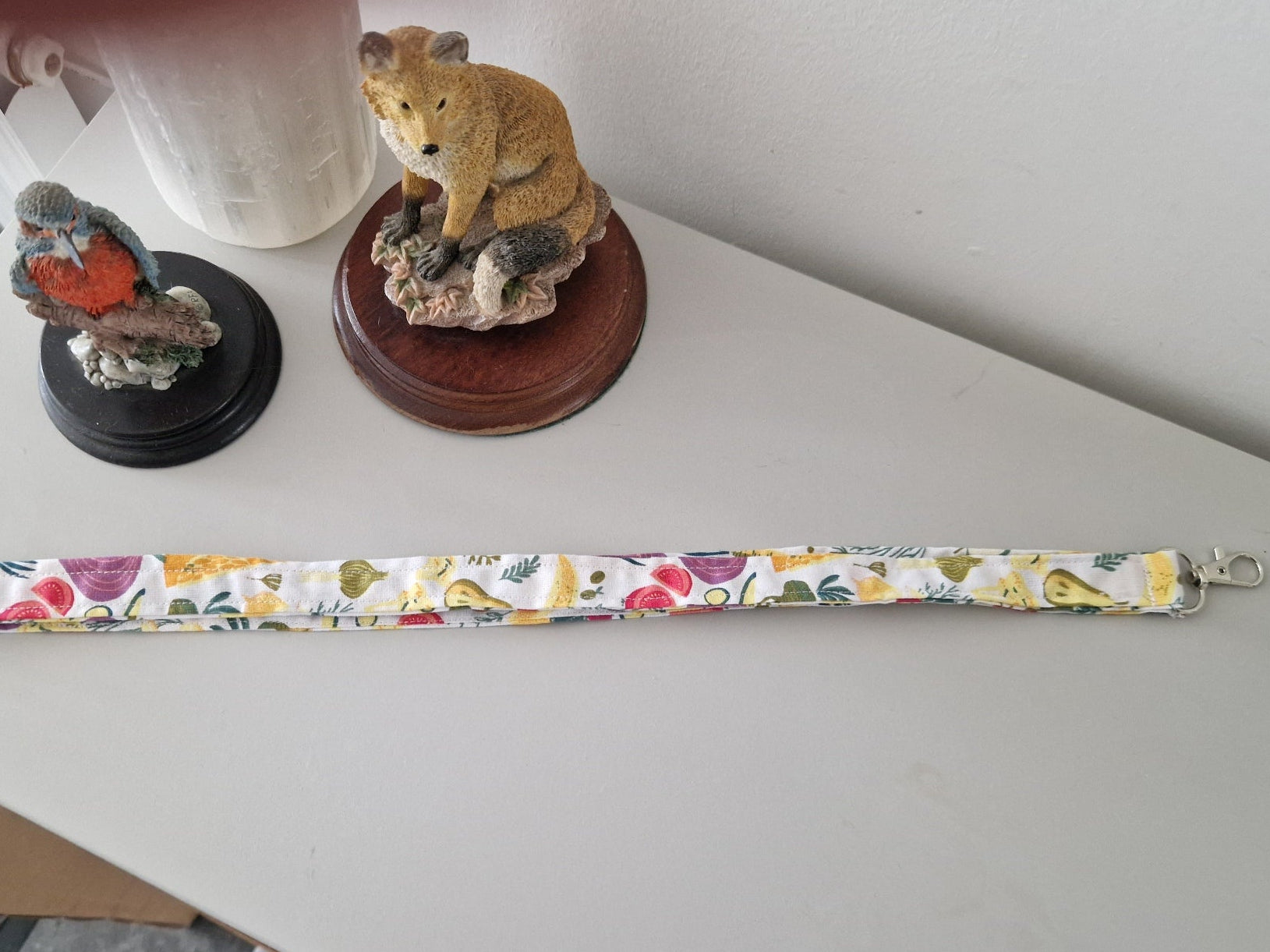 A fabric lanyard  strap with a veg pattern, laid out on a light gray tabletop. The strap has silver metal clasps on both ends and is photographed in a home setting with decorative figurines in the background.
