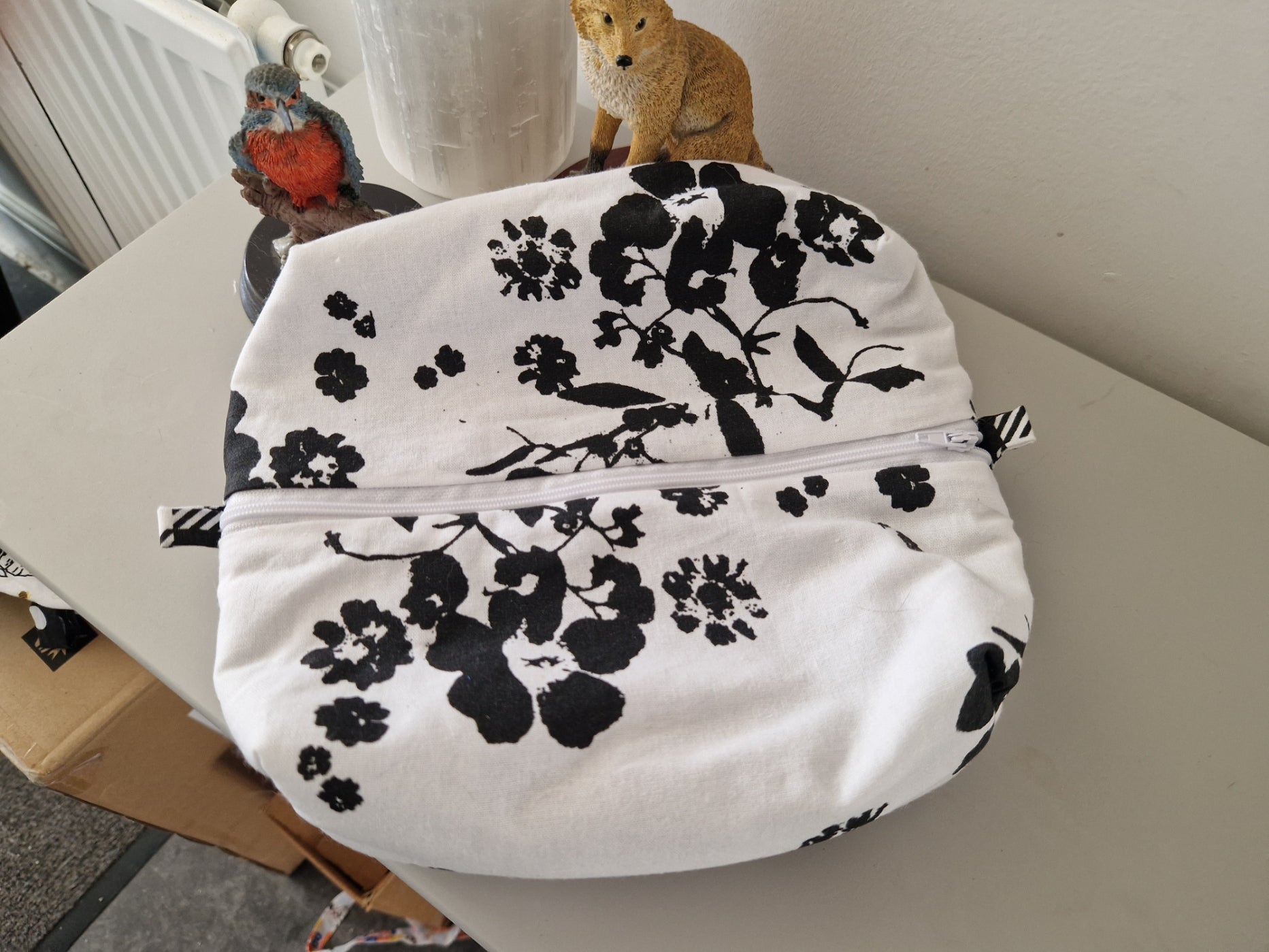 A white fabric zippered pouch with a black floral print, shown on a tabletop with decorative figurines in the background.