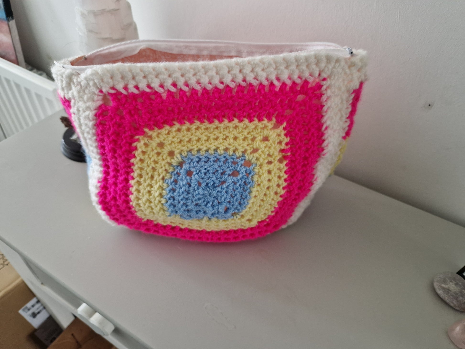 A handmade crochet makeup bag featuring a bright granny square pattern with concentric squares of light blue, yellow, hot pink, and white wool, featuring a white top zipper closure.