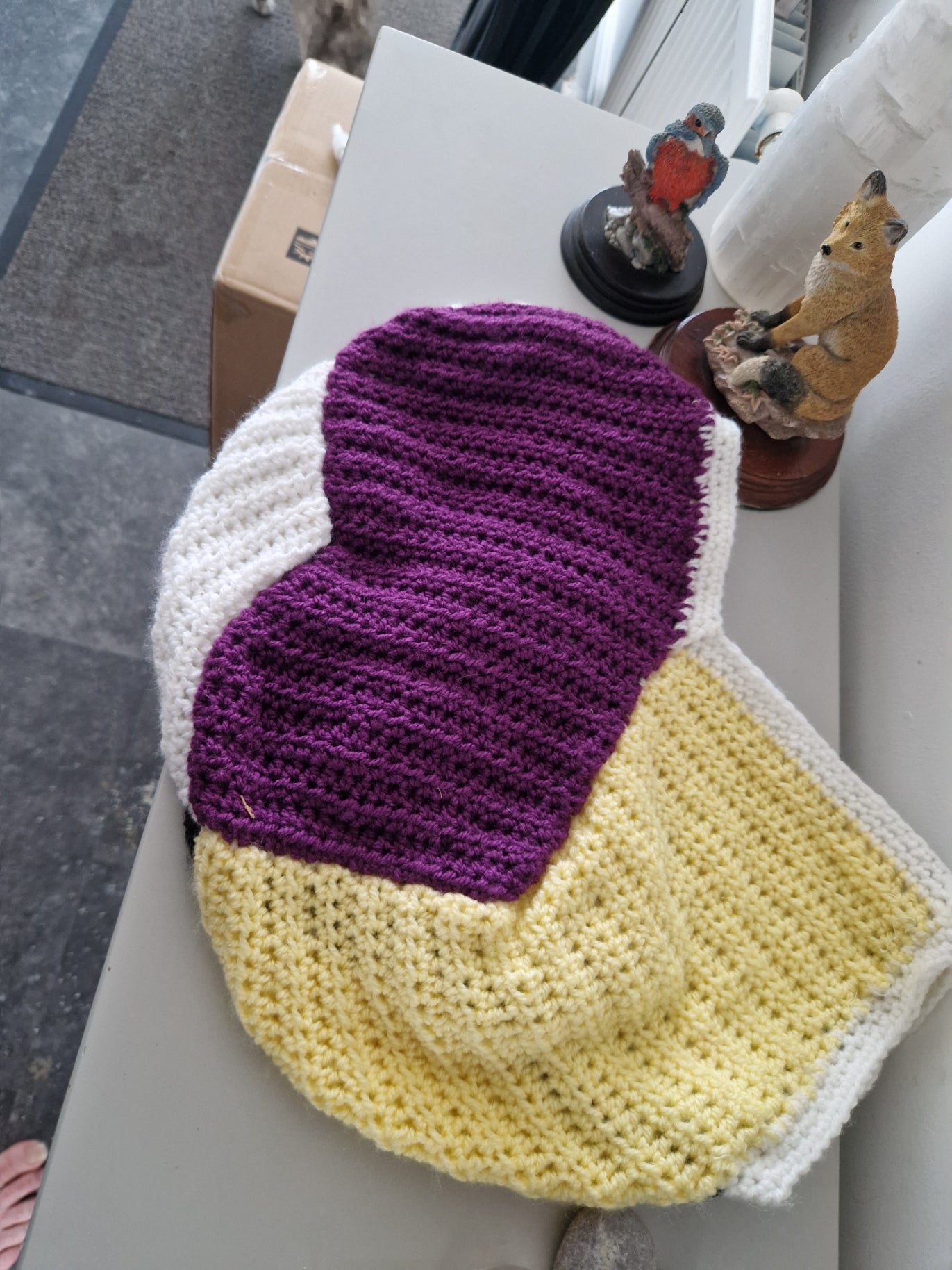 A crocheted stocking with three main color sections: purple, yellow, and white, displayed on a light gray table with decorative figurines.