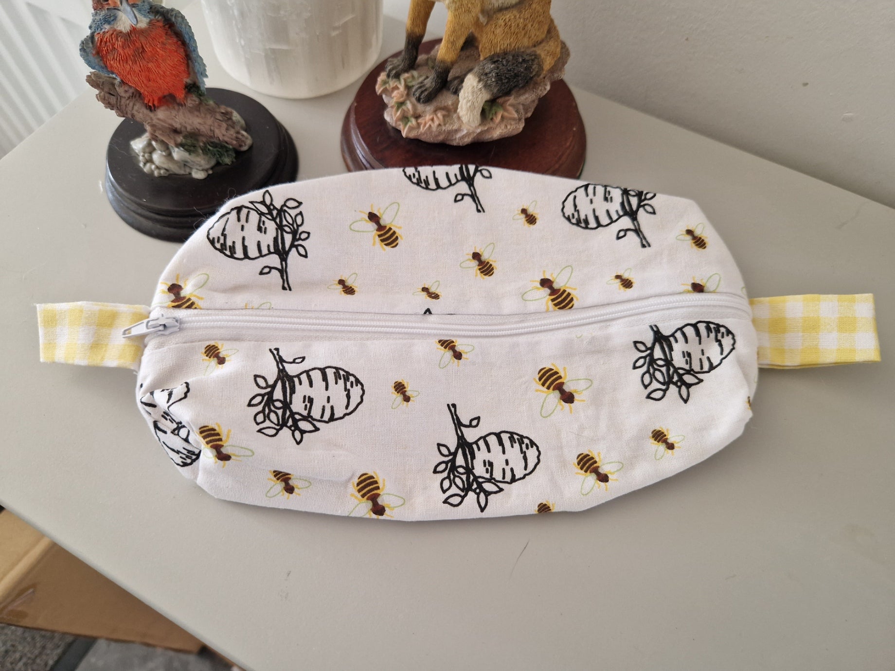 A white zippered pouch with a black bee and honeycomb print, featuring yellow-and-white gingham side straps, resting on a light gray tabletop.