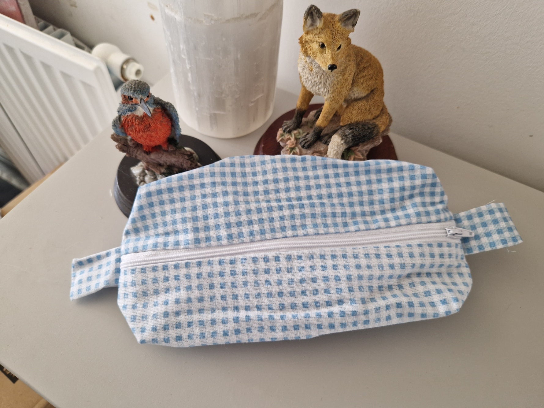 A light blue and white gingham zippered pouch with a fabric loop strap, resting on a gray tabletop in a home setting with decorative bird and fox figurines in the background.