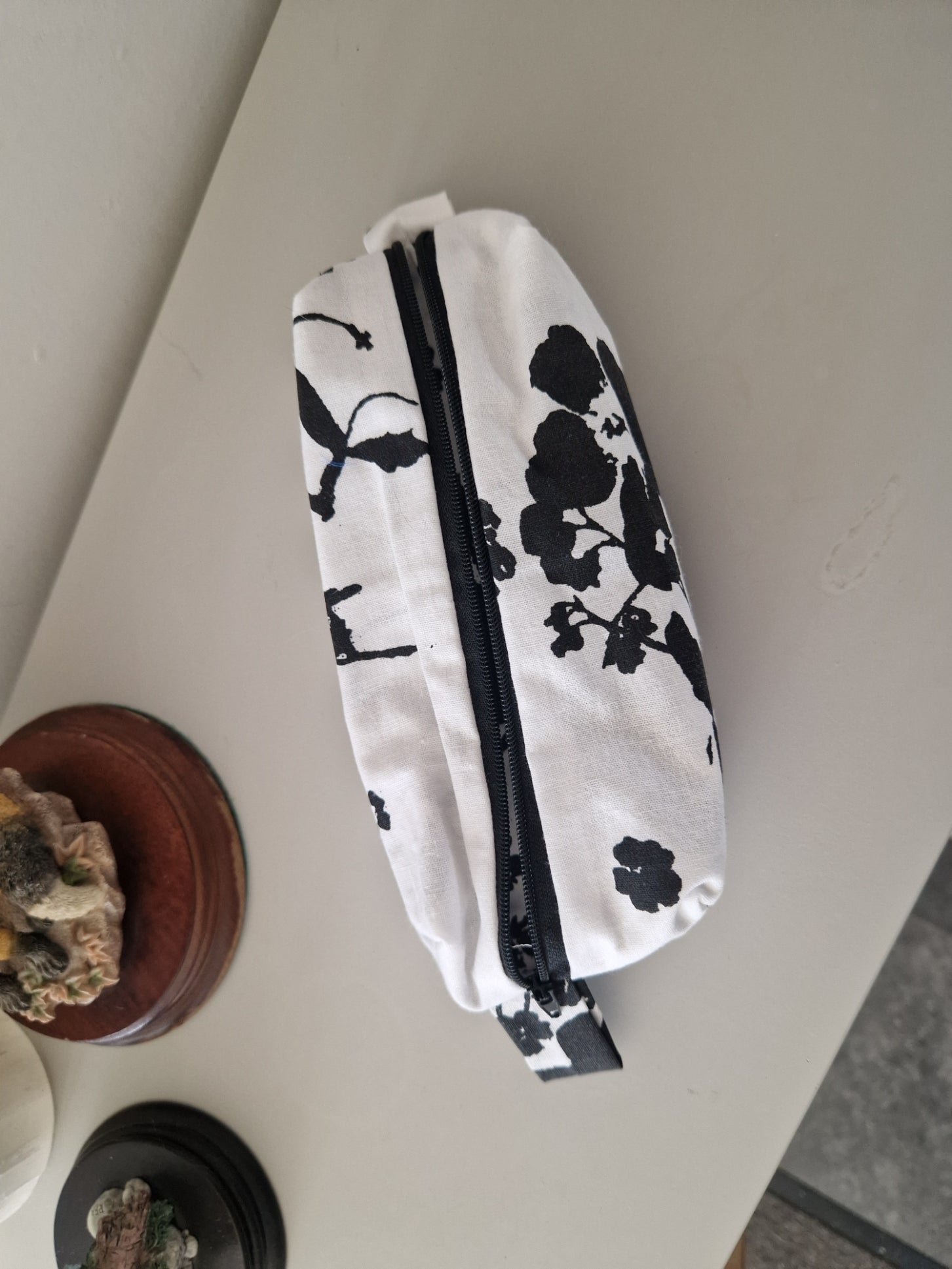A white fabric zippered pouch with a black floral print, resting on a light gray tabletop.