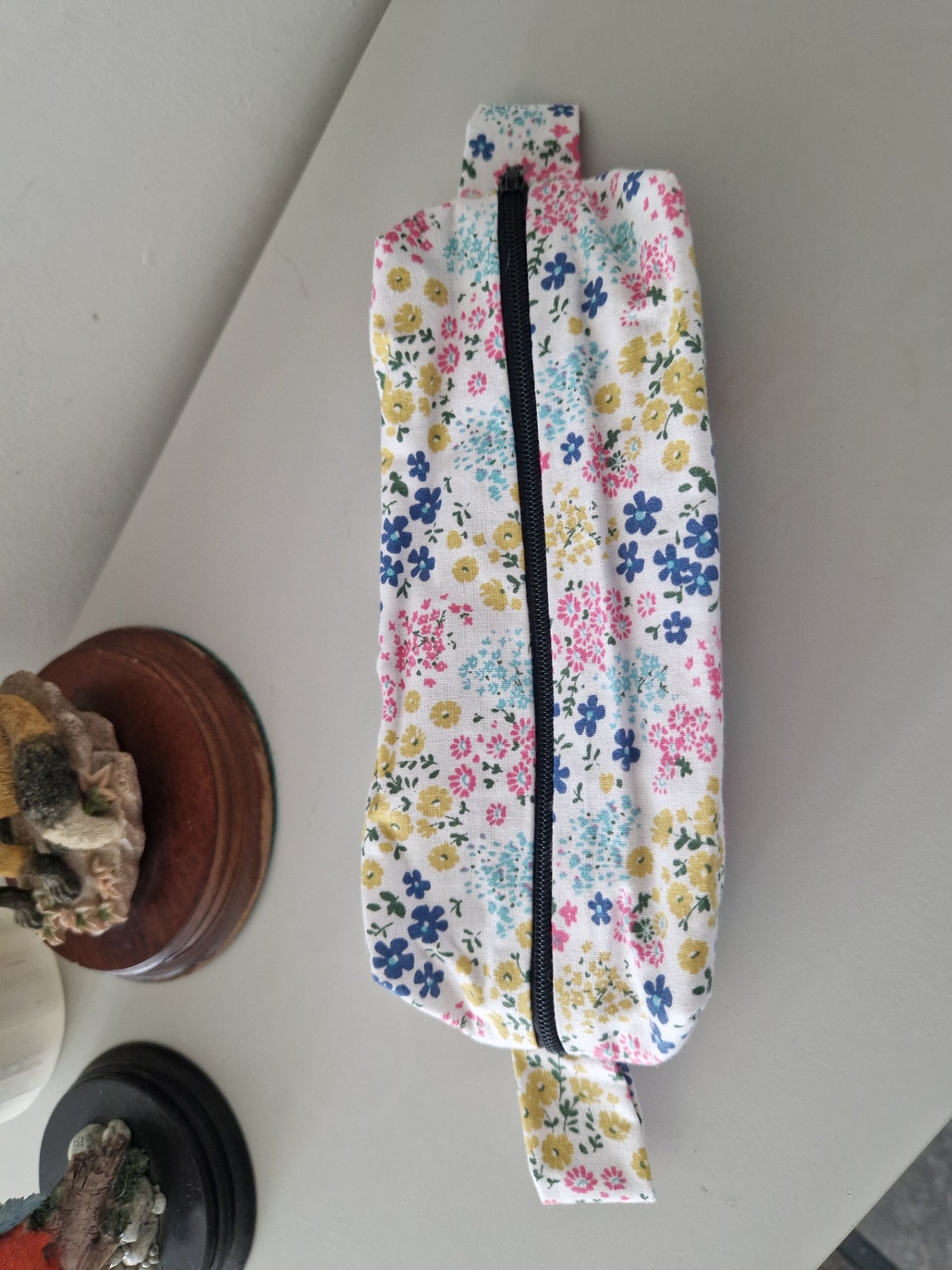 A fabric zippered pouch with a colorful floral print on a white background, featuring a black zipper and a small side loop, resting on a light-colored tabletop.