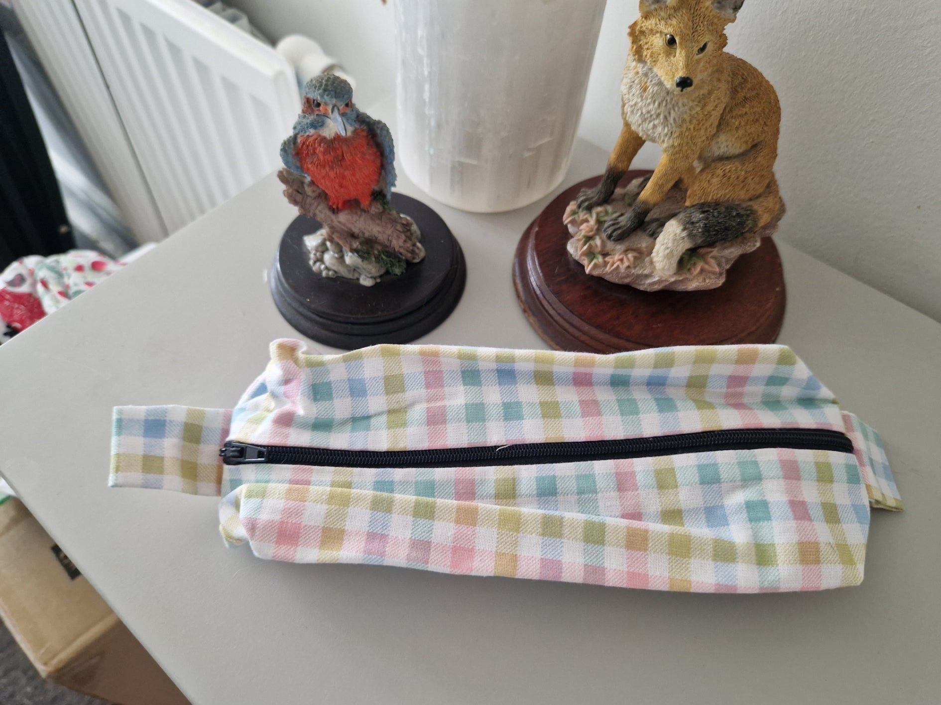 A pastel plaid zippered pouch with a black zipper, placed on a gray tabletop in front of two decorative figurines.