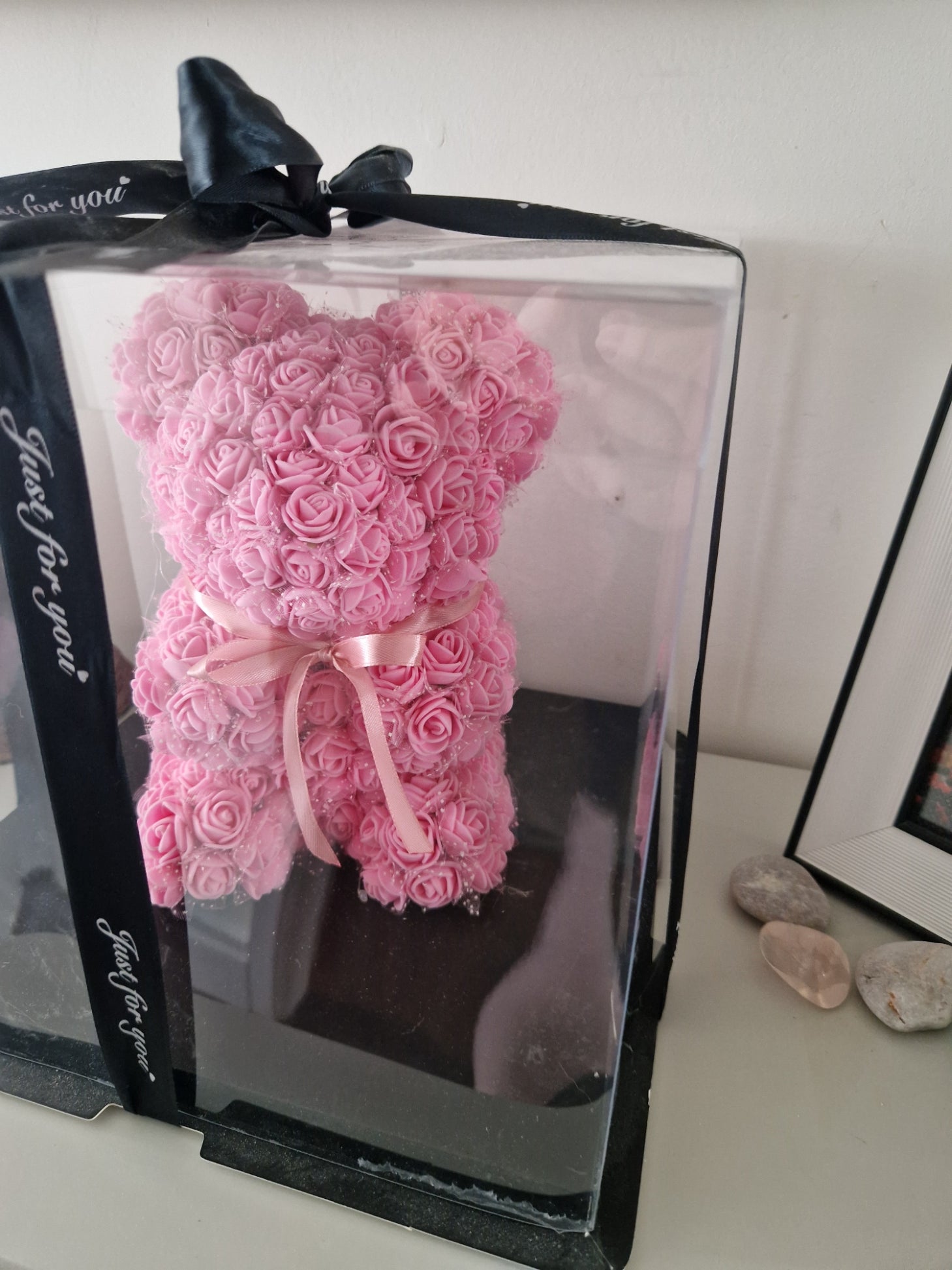 A pink rose-shaped teddy bear gift box with a clear plastic lid, black ribbon bows, and 'Just for you' printed on the black ribbon, photographed on a light surface.