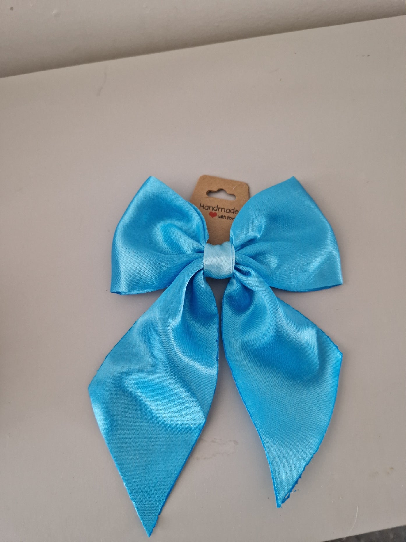 A light blue satin hair bow with a small cardboard tag attached, placed on a plain beige surface.