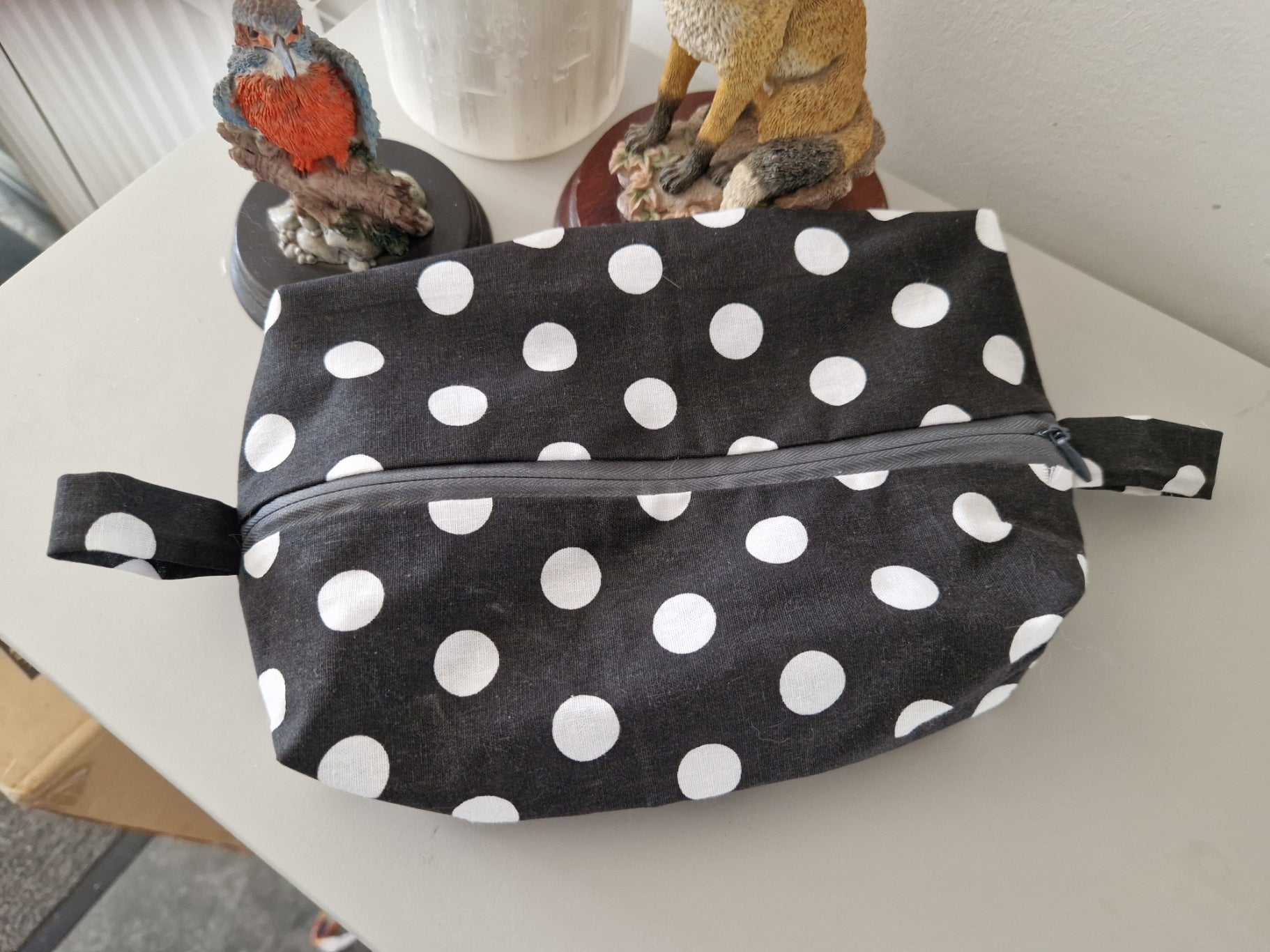 A black fabric zippered pouch with white polka dots, resting on a light-colored tabletop with decorative figurines in the background.