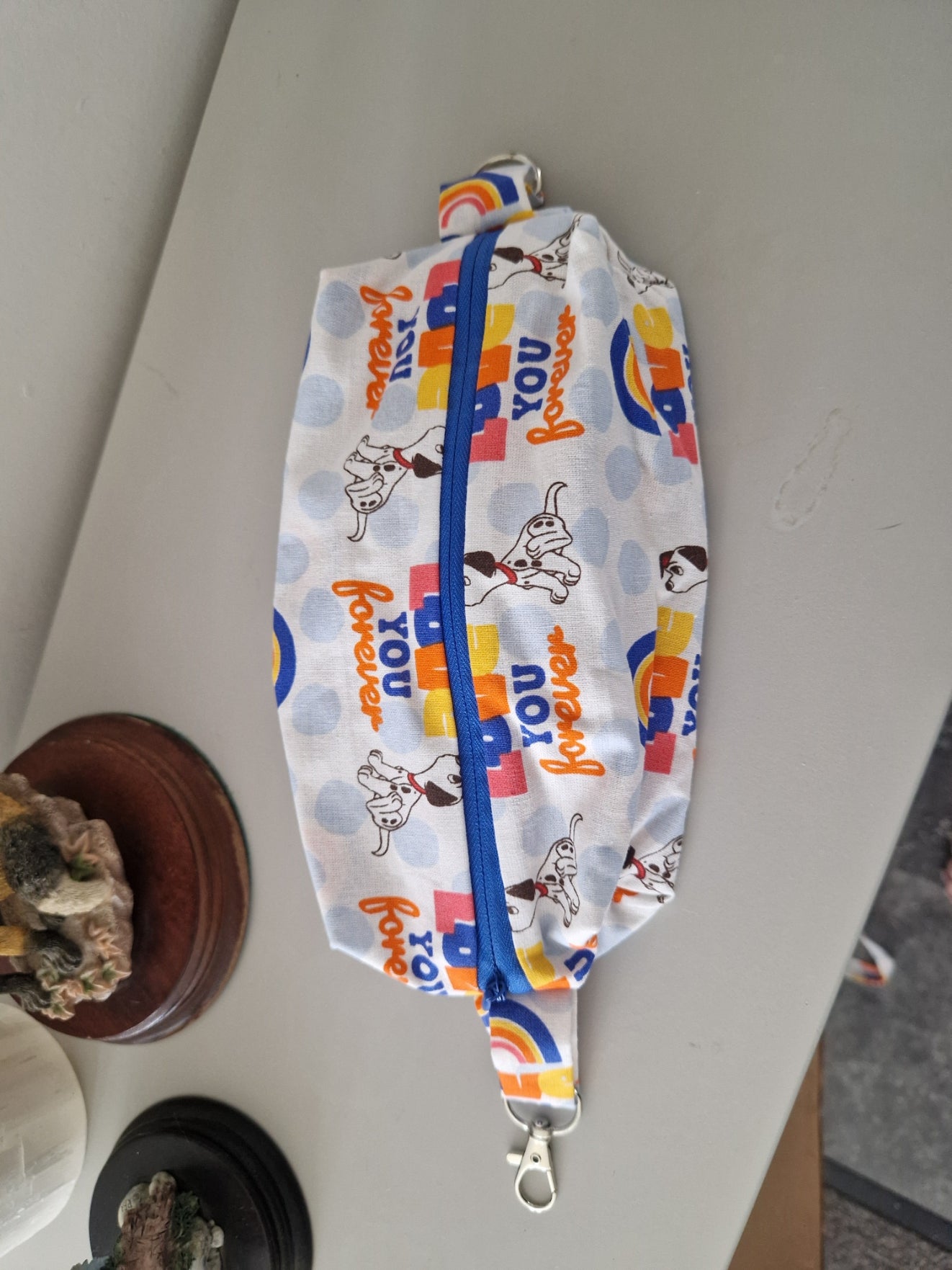 A fabric waist bag with a white background, blue polka dots, and a colorful cartoon dog print with the words 'you are so cool'. It has a blue zipper, silver metal clips on both ends, and is photographed on a light gray tabletop.