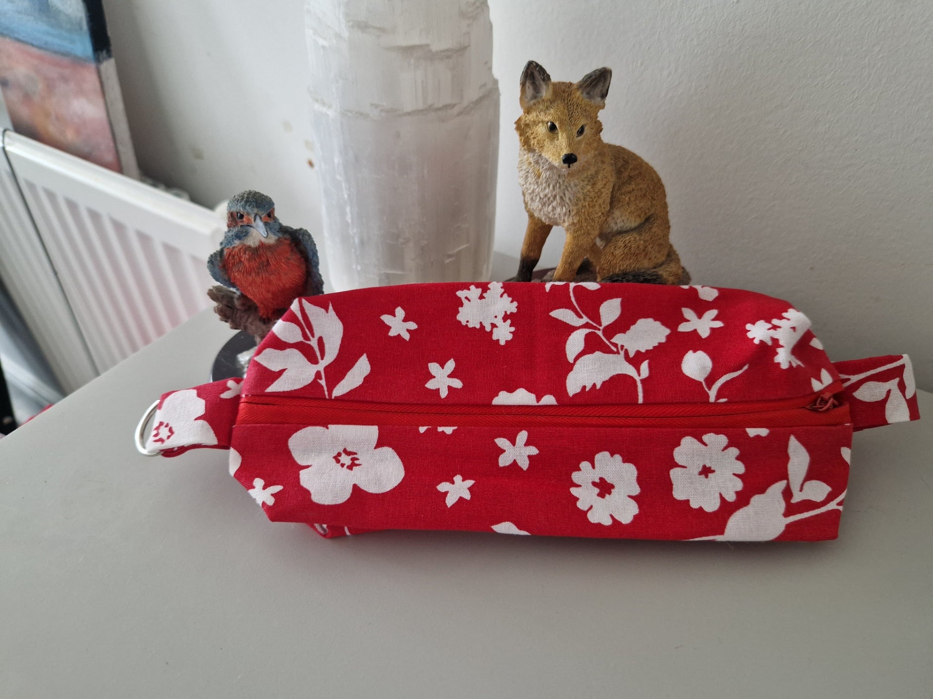 A red fabric zippered pouch with a white floral print, resting on a gray tabletop with decorative figurines in the background.