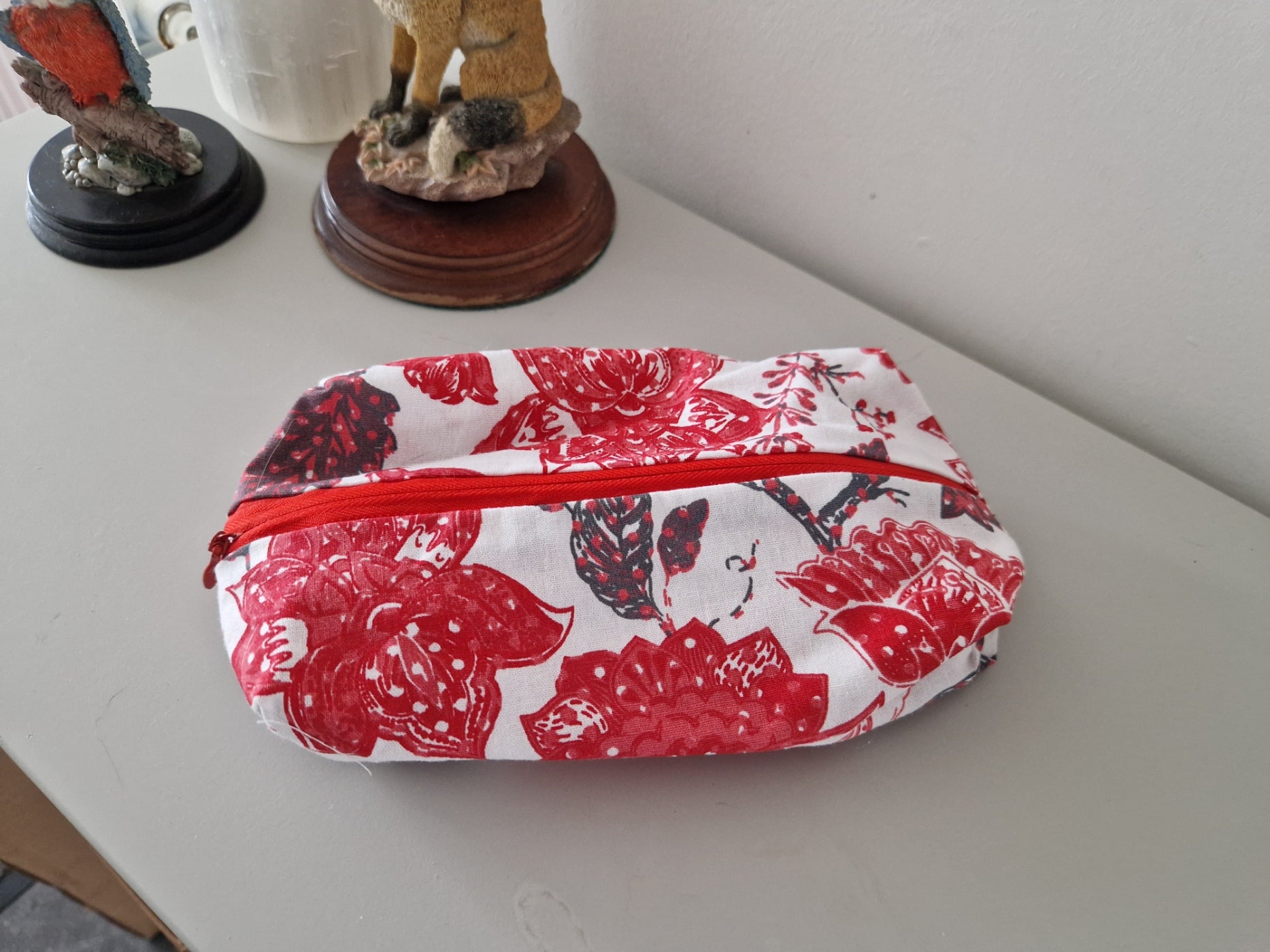 A rectangular zippered pouch with a red and white floral print sits on a light gray tabletop, with small decorative figurines in the background.